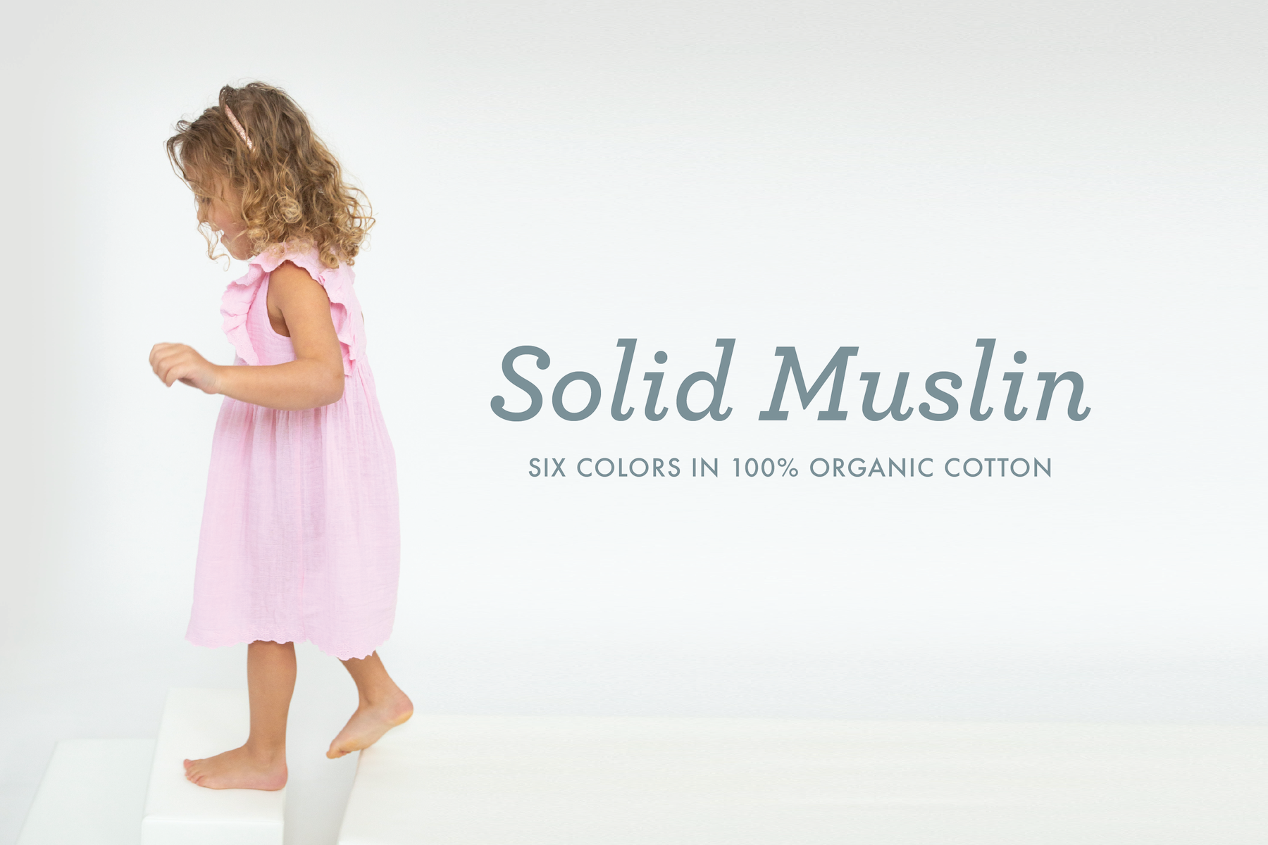girl with curly hair in a pink dress walking on white block. Text says Solid Muslin with six colors in 100% organic cotton