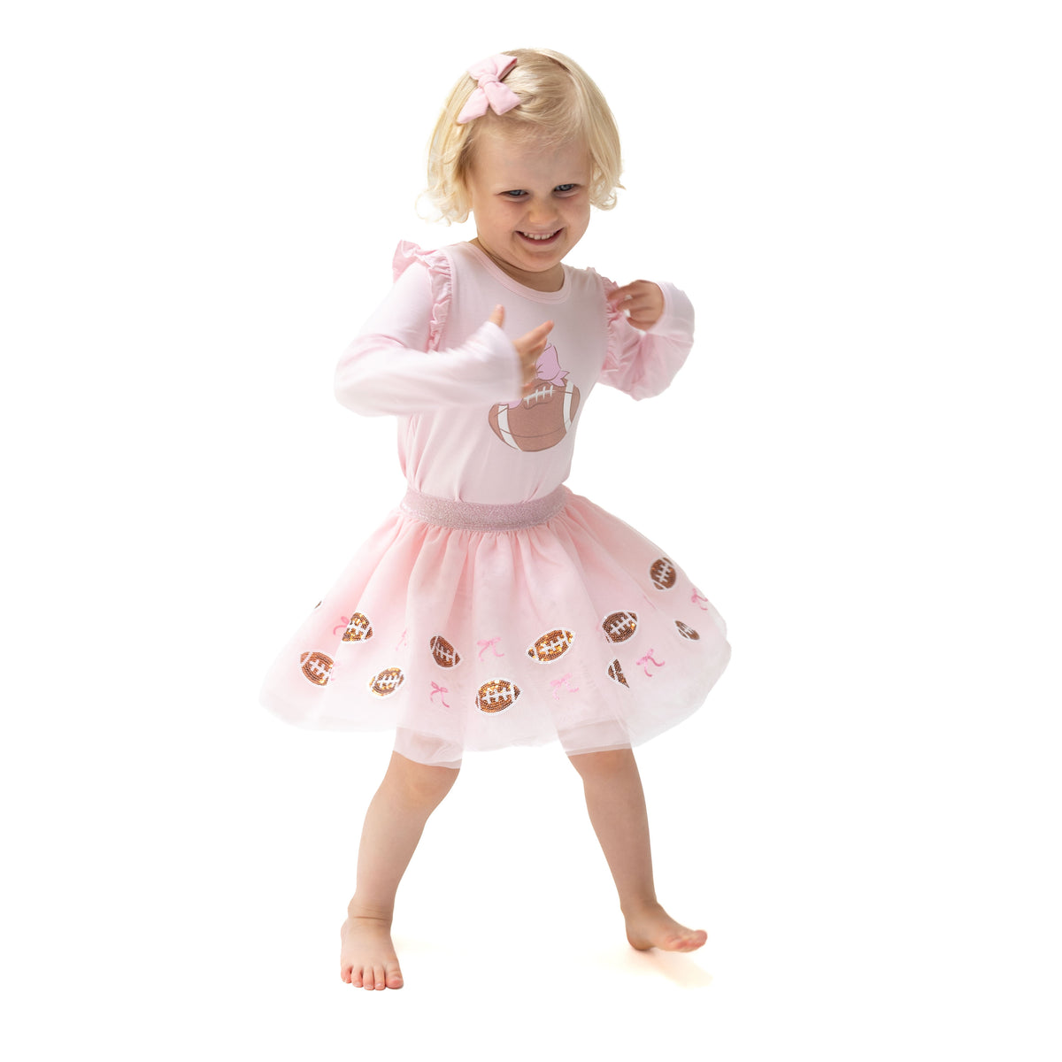 A young toddler girl wearing a pink long-sleeve tulle dress with football graphics on the skirt, posing on a solid white background.
