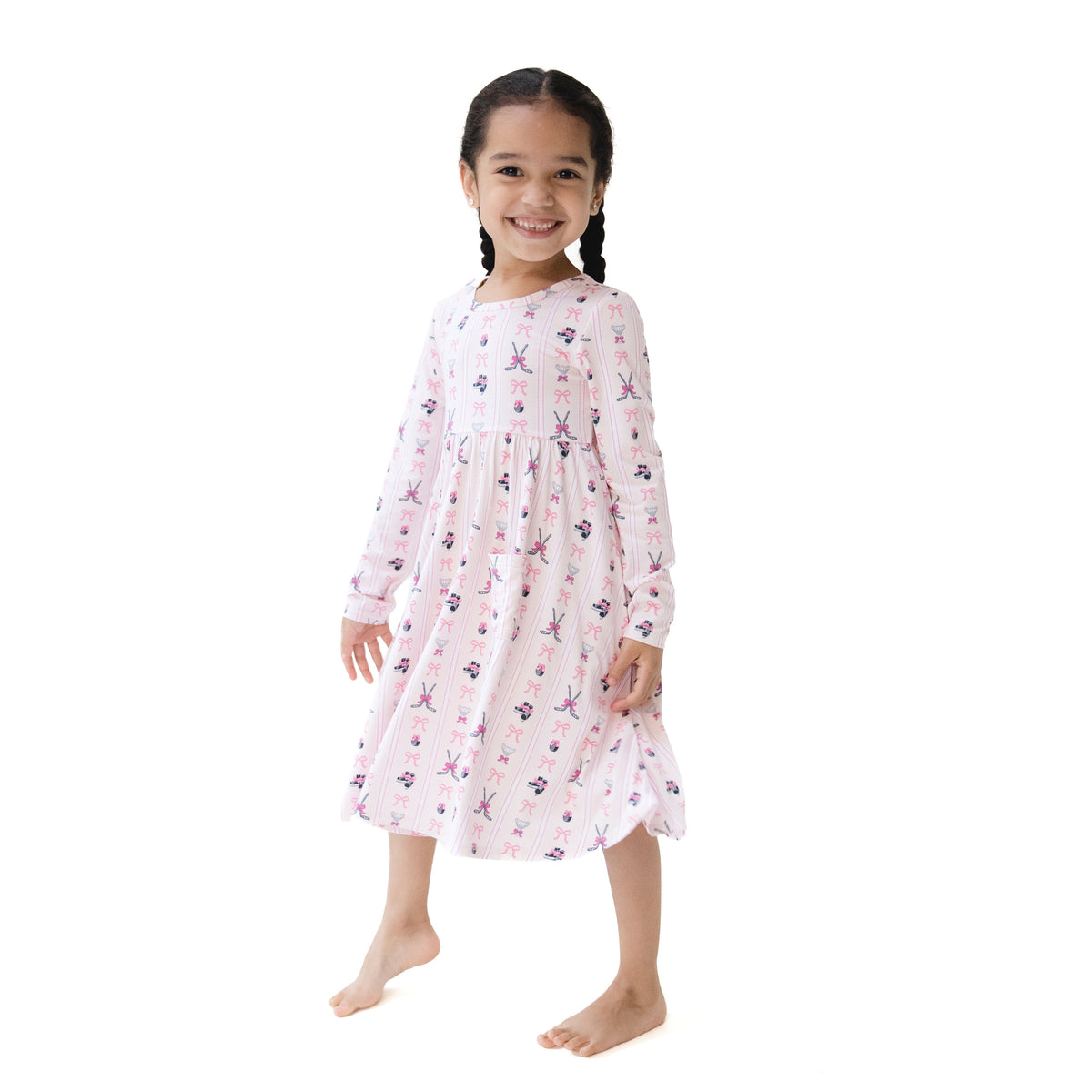 A young girl with braided hair stands barefoot against a plain white background, wearing a long-sleeve baby dress with a pink and white pattern of hockey stick and bows.