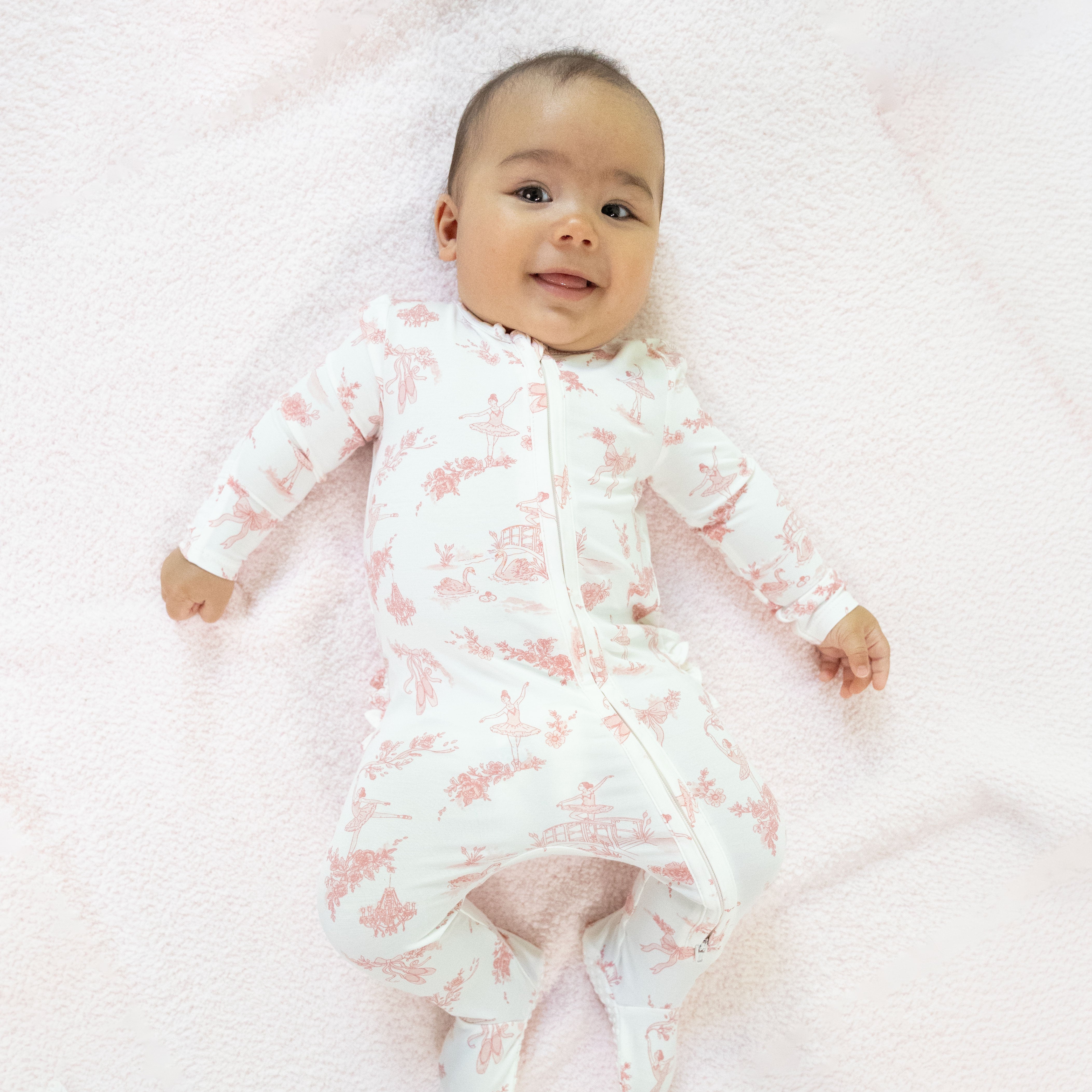 A baby lying on a soft pink blanket, wearing a white long-sleeve footless sleeper with a pink toile-style print and a front zipper.