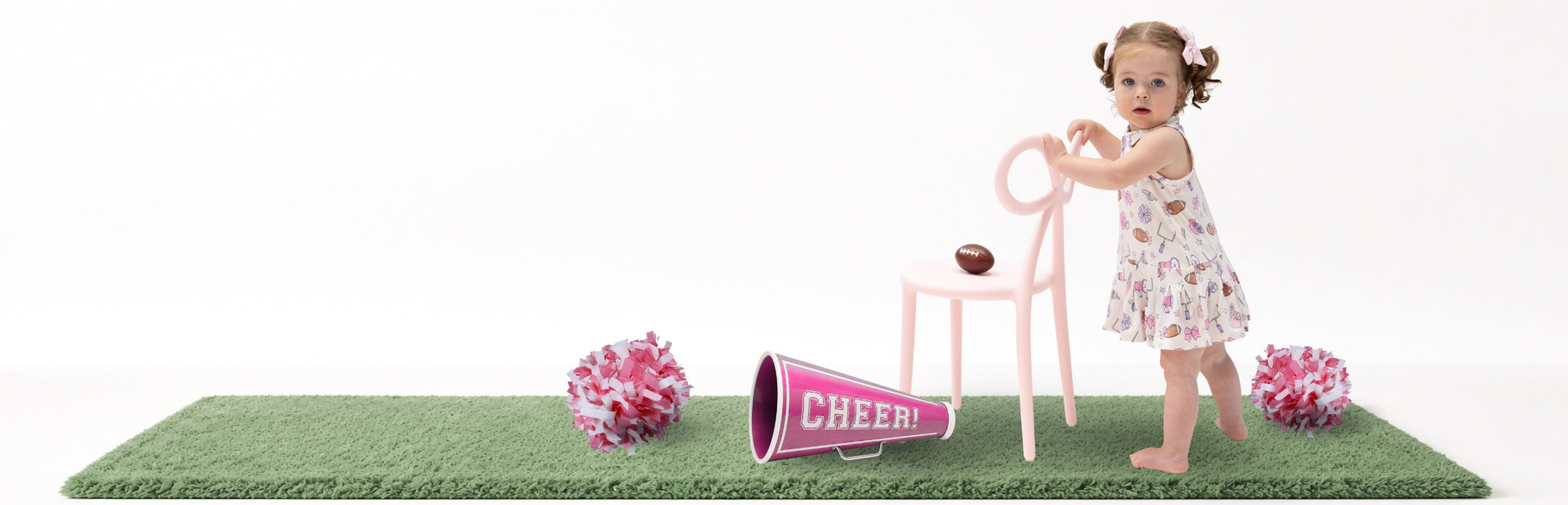 A toddler girl in a football  cheerleader dress stands next to a pink chair and megaphone on a green mat, with pink pom-poms on a white background.