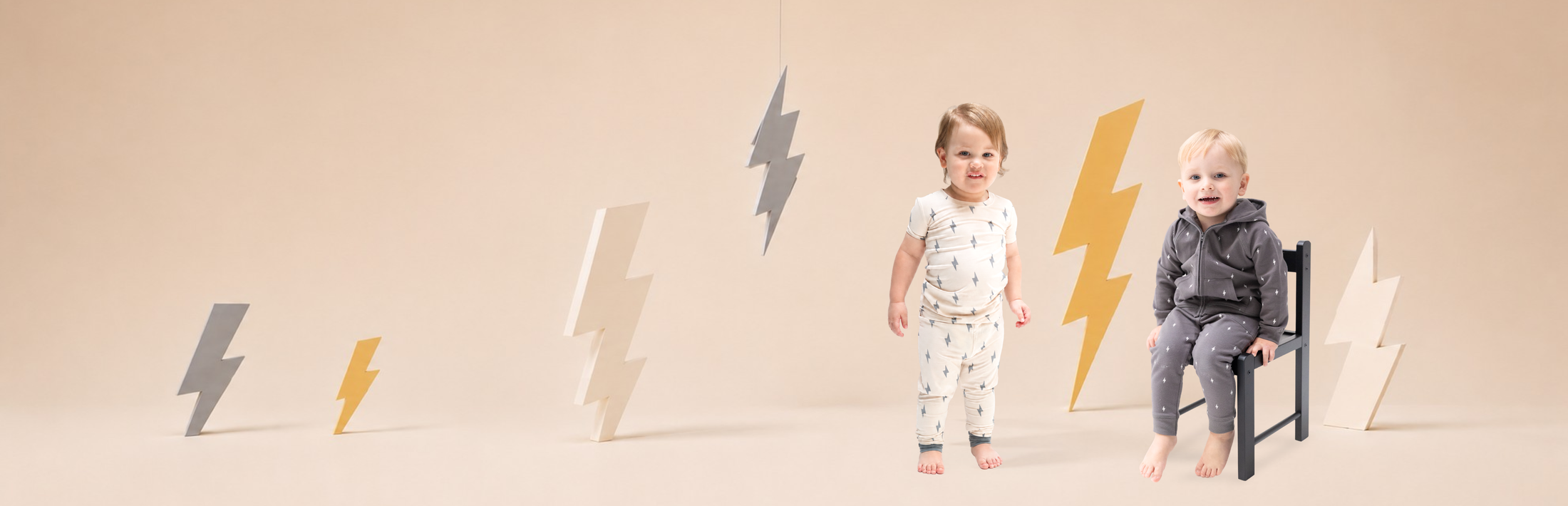 Two toddler boys wear lightening bolt printed outfits on a warm white background with large lightening bolt props around the set.