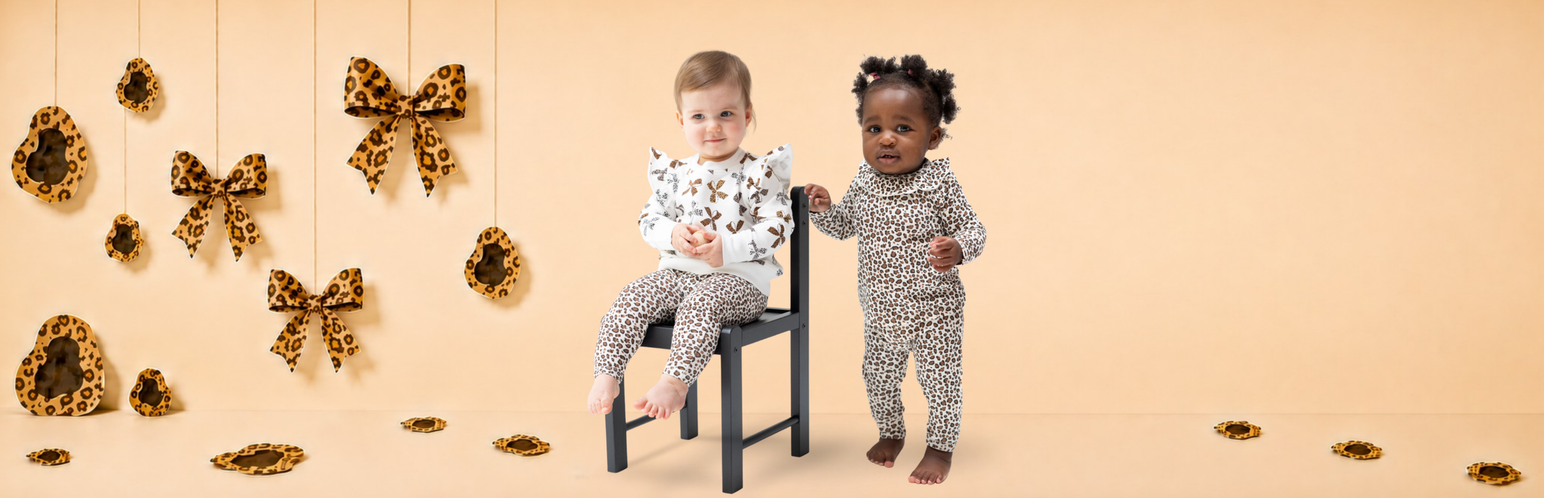 Two toddlers modeling leopard-print outfits. One wears an oversized sweat shirt and leggings and the other wears a one piece with stylized collar. The studio setting is a beige backdrop, hanging leopard bows and spots, and a small black chair.