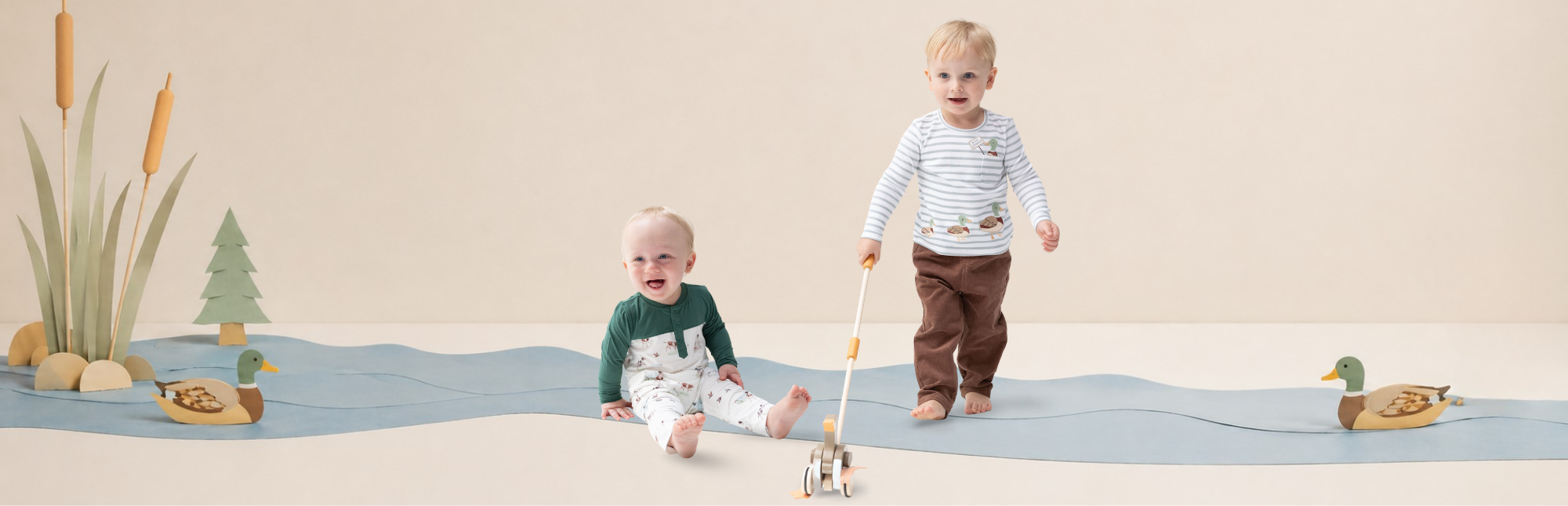 Two toddlers wearing outfits from the duck collection. One toddler stands holding a small toy boat, wearing a long sleeved striped shirt with embroidered duck appliques and brown corduroy pants. The other toddler (sitting) wears a romper with a dog and duck print. The set is a stylized wooden river scene made with large paper crafts.
