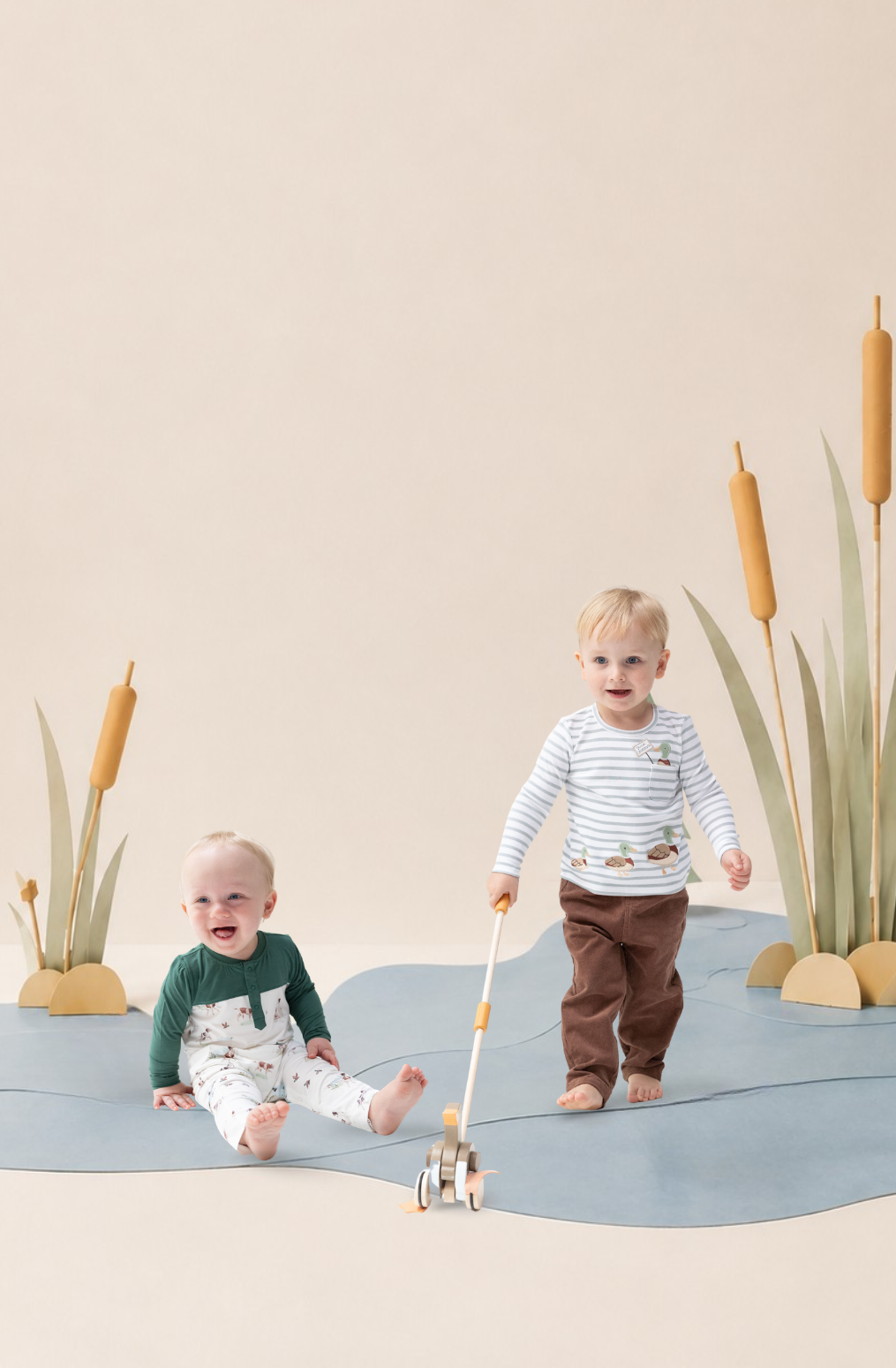 Two toddlers wearing outfits from the duck collection. One toddler stands holding a small toy boat, wearing a long sleeved striped shirt with embroidered duck appliques and brown corduroy pants. The other toddler (sitting) wears a romper with a dog and duck print. The set is a stylized wooden river scene made with large paper crafts.