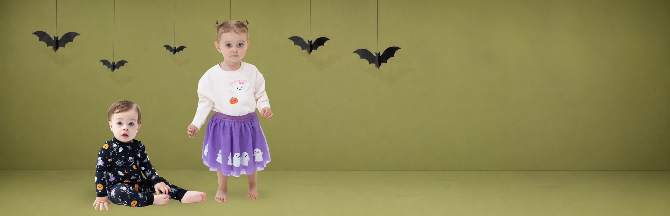 A toddler boy in dark glow-in-the-dark Halloween pajamas  and girl toddler wearing a purple tutu with white sequined ghosts and a light french terry sweat shirt with a ghost and pumpkin in a studio with an olive green background and hanging black bat decorations.