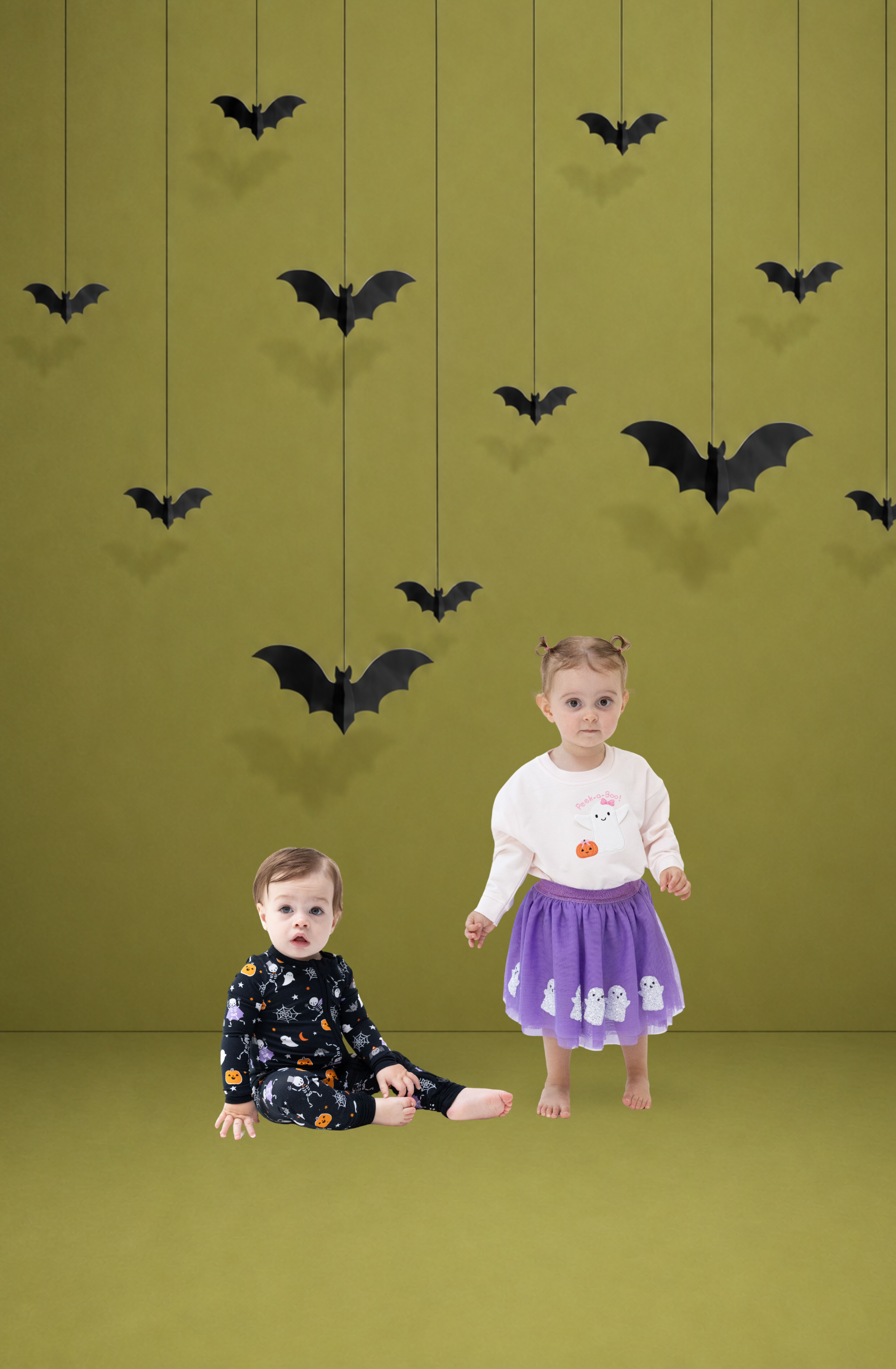 A toddler boy in dark glow-in-the-dark Halloween pajamas  and girl toddler wearing a purple tutu with white sequined ghosts and a light french terry sweat shirt with a ghost and pumpkin in a studio with an olive green background and hanging black bat decorations.
