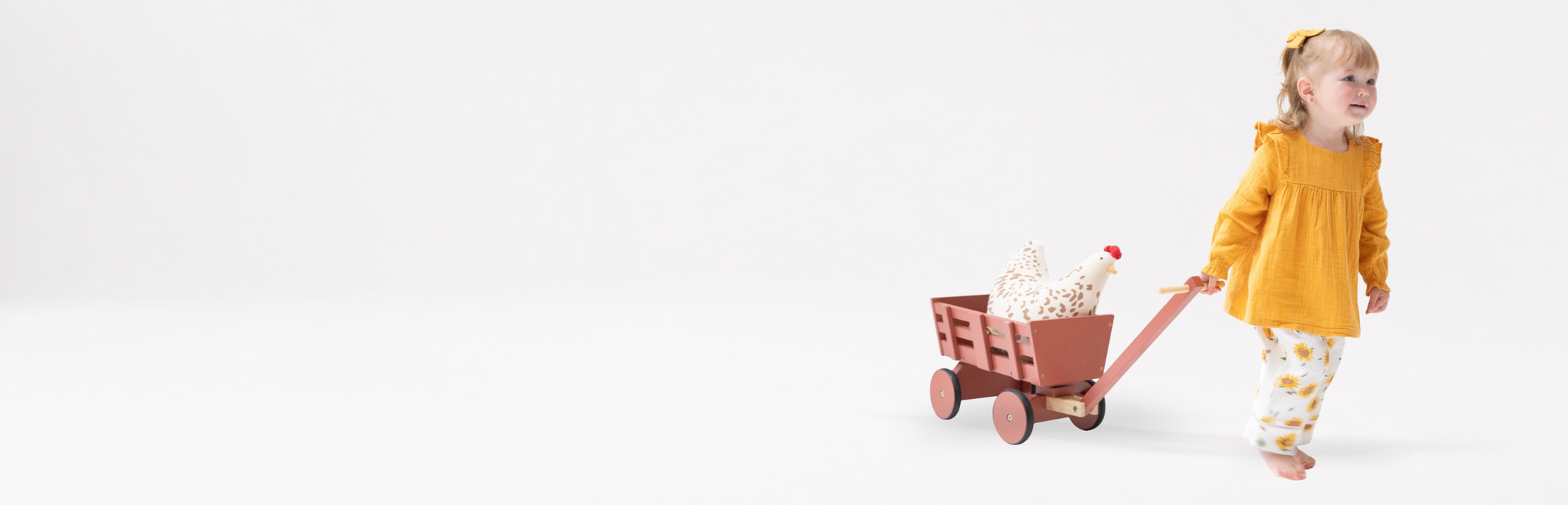 A toddler girl in a muslin bright yellow long-sleeve top and muslin yellow-and-white patterned pants pulls a small wooden wagon with a white stuffed animal chicken, against a seamless white studio background.