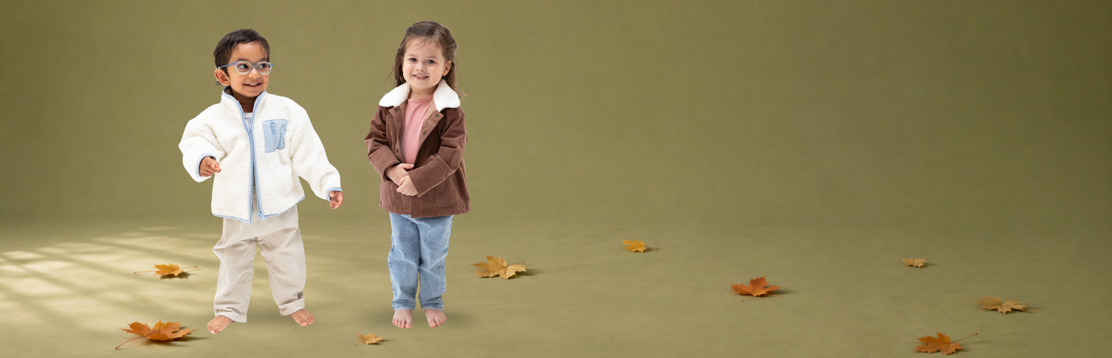 Two young children standing barefoot on a solid olive green studio floor, surrounded by scattered autumn leaves. The child on the left wears a white chenille jacket with a light blue chest panel and beige pants, while the child on the right wears a brown corduroy jacket with a white sherpa collar, a pink top, and blue jeans.