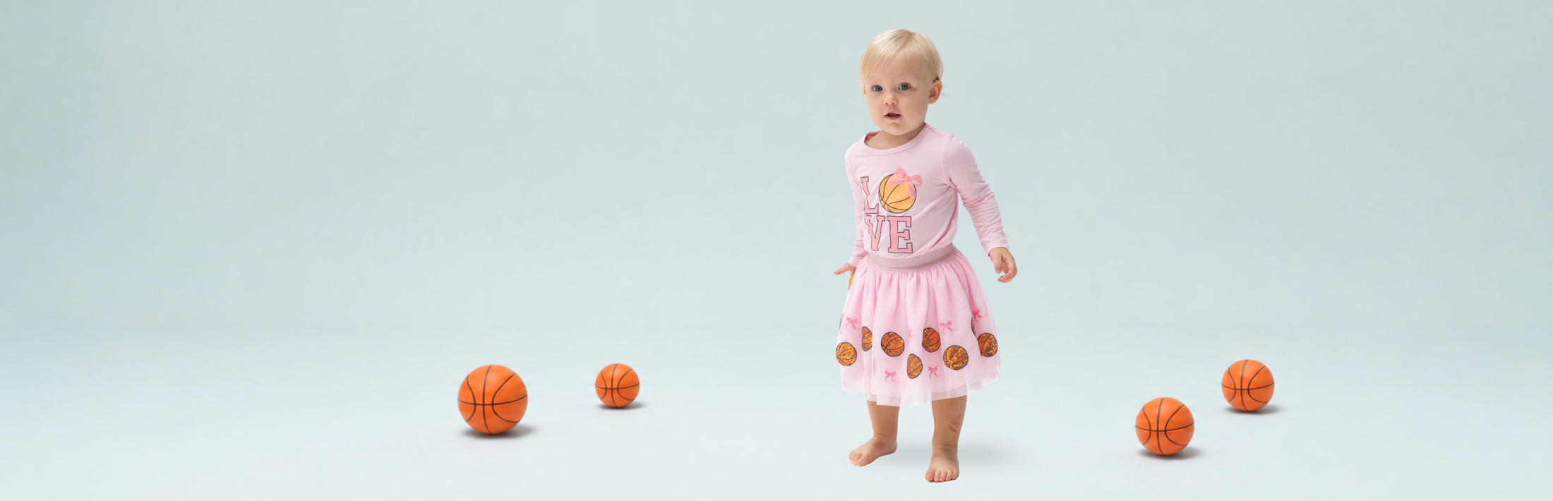A toddler girls stands barefoot in a light blue studio setting wearing a pink long-sleeve shirt with basketball graphics, and a basketball tutu skirt surrounded by several small orange basketballs.