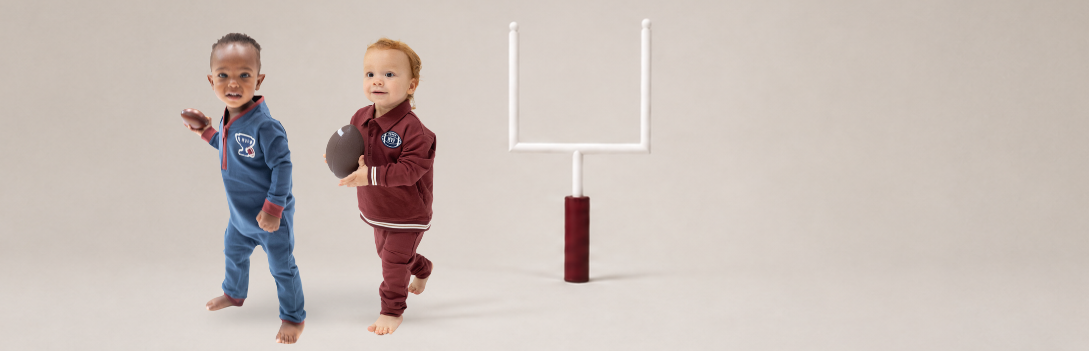Two toddler boys in football-themed organic french terry tracksuits, one in blue and one in maroon, standing next to a miniature white football goal on a neutral studio background.