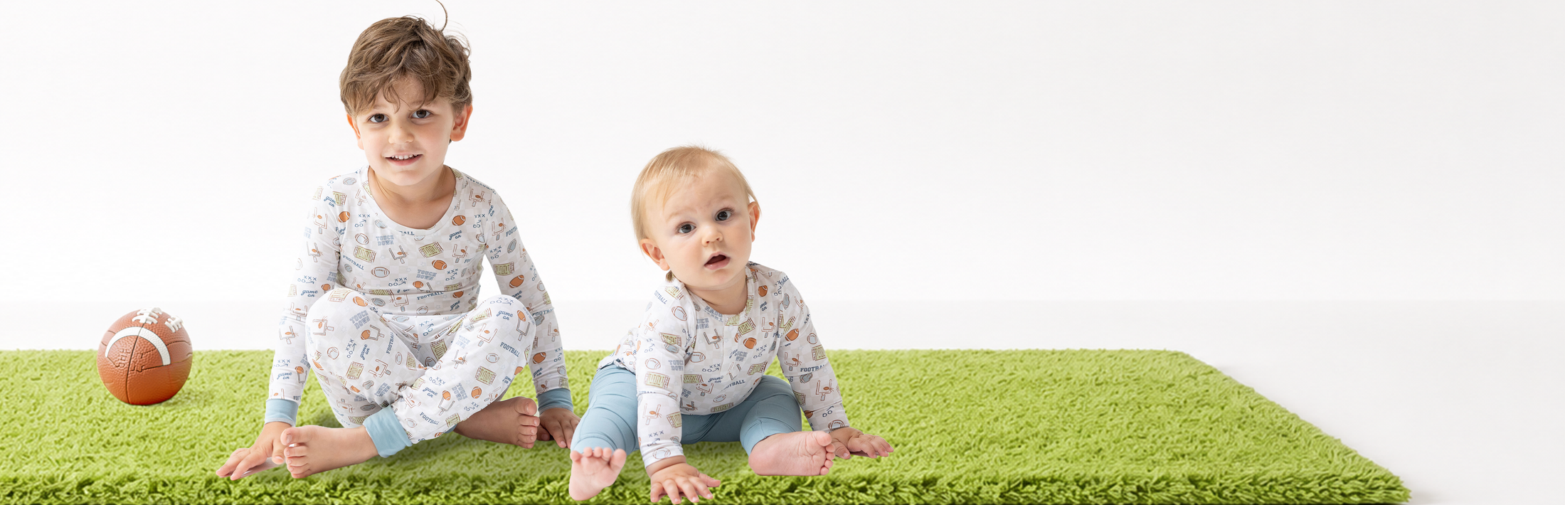 A baby boy and older older brother wearing football printed outfits in soft bamboo fabric, sitting on a bright green rug with a football, against a white studio background.