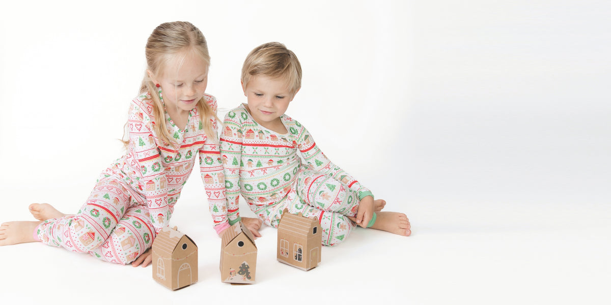Two children sitting on a white floor wearing matching holiday-themed long-sleeve pajamas with a green, red, and white pattern, looking at small kraft paper gingerbread houses toys.