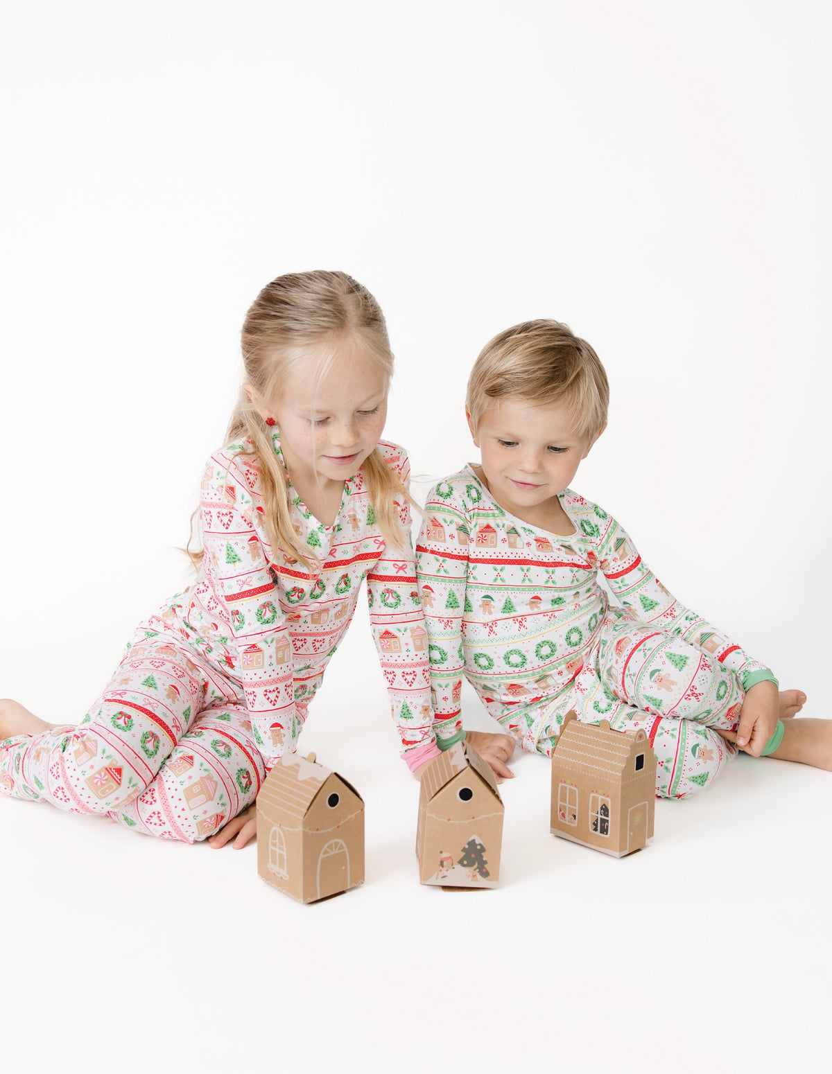 Two children sitting on a white floor wearing matching holiday-themed long-sleeve pajamas with a green, red, and white pattern, looking at small kraft paper gingerbread houses toys.