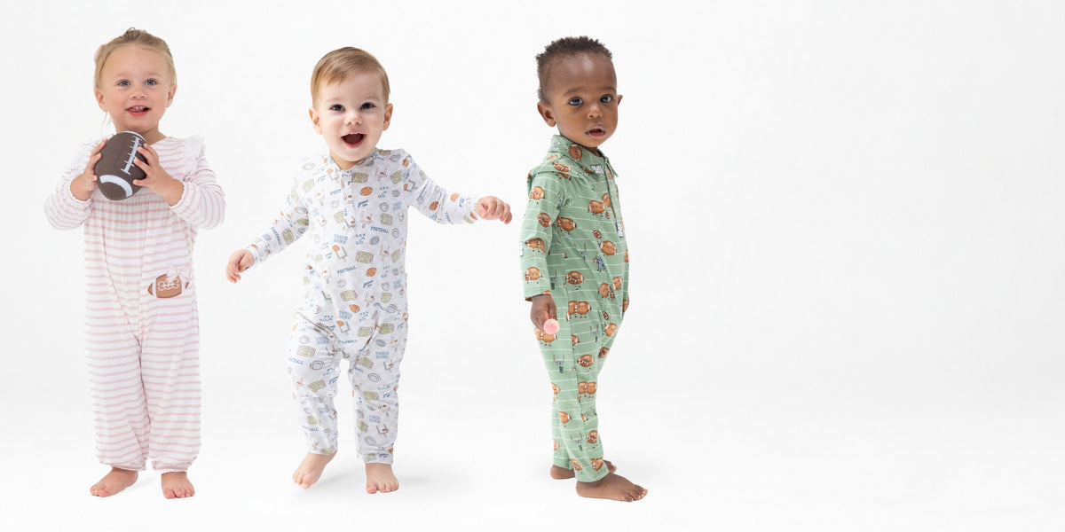 Three toddlers standing side by side in football themed patterned play rompers against a clean white studio background. The child on the left wears pale pink and white stripes and holds a small football.