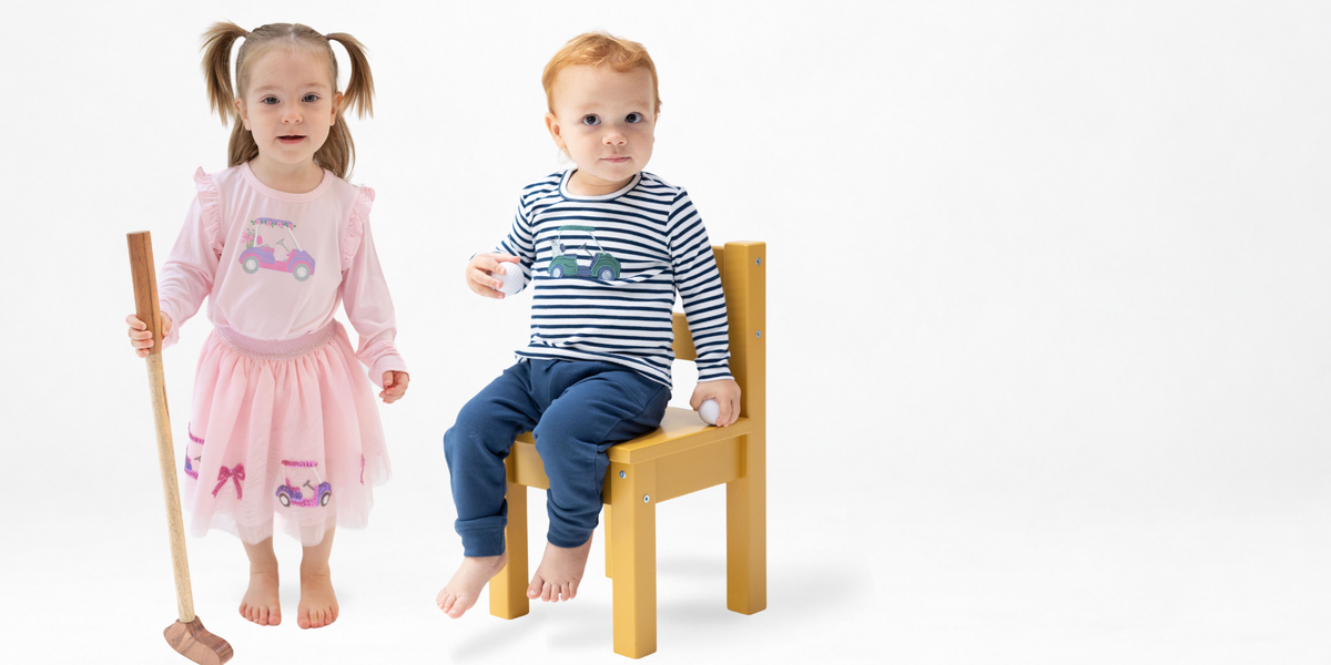 Girl and boy toddler wear golf play wear. The girl is wearing a long sleeved pink shirt with ruffles at the shoulder and a golf cart on the chest paired with a golf tutu. The boy wears a classic navy and white striped long sleeve shirt with a green golf cart applique on the chest paired with navy french terry jogger pants.