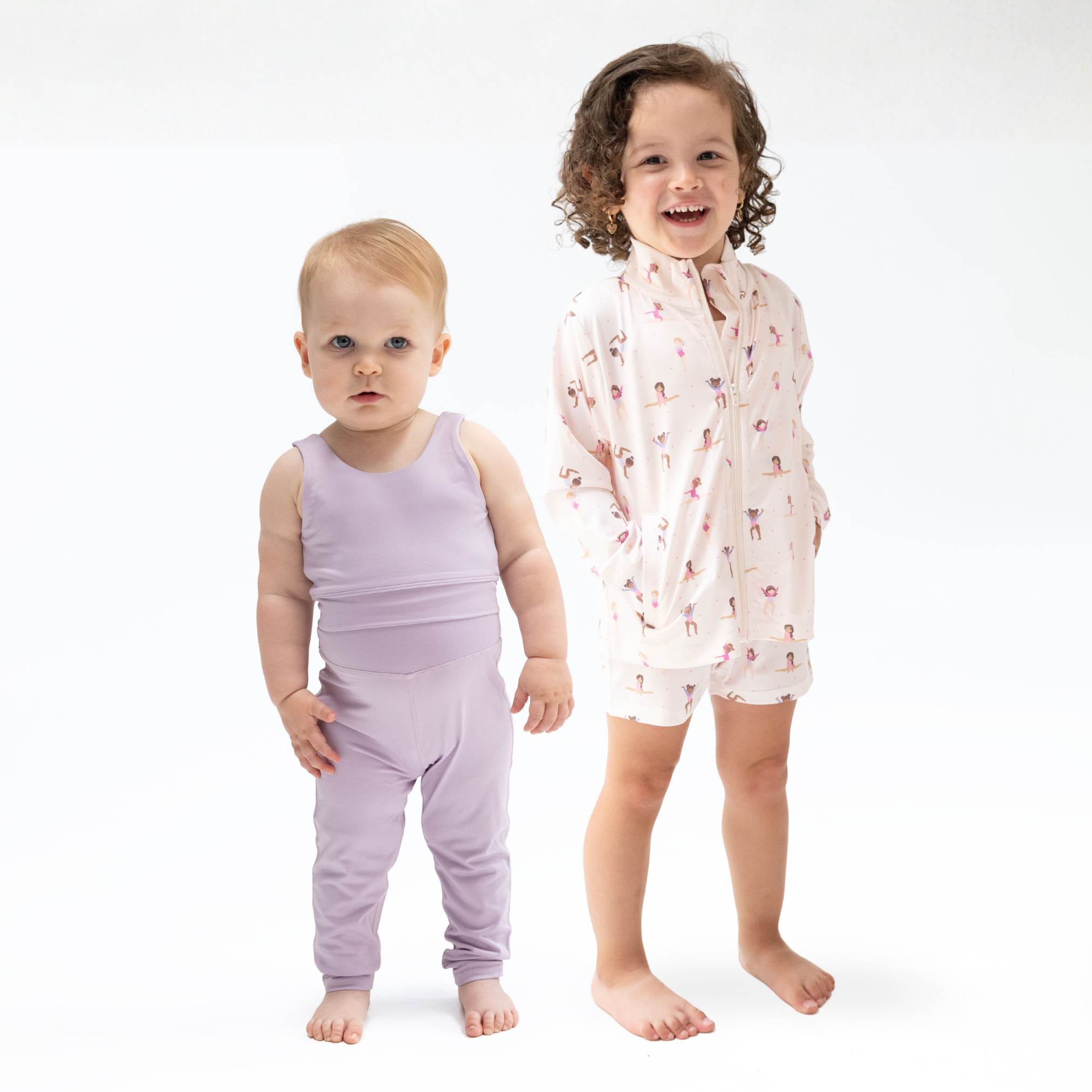 Two young children standing barefoot against a clean white studio background. The toddler on the left wears a solid light lavender sleeveless two-piece activewear outfit, while the older child on the right wears a patterned long-sleeve two piece activewear outfit.