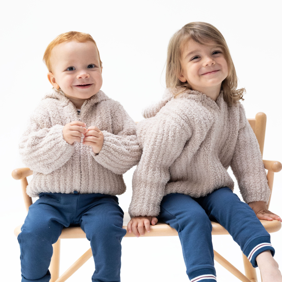 Two toddlers sitting on a wooden chair against a white background, wearing matching beige sherpa zip-up hoodies and blue pants.
