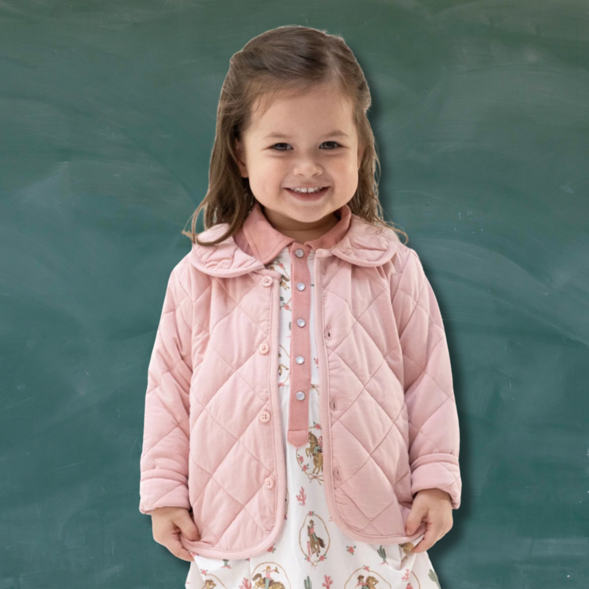 A young girl smiling and wearing a light pink quilted jacket over a white dress with a floral western print, standing in front of a dark green chalkboard background.
