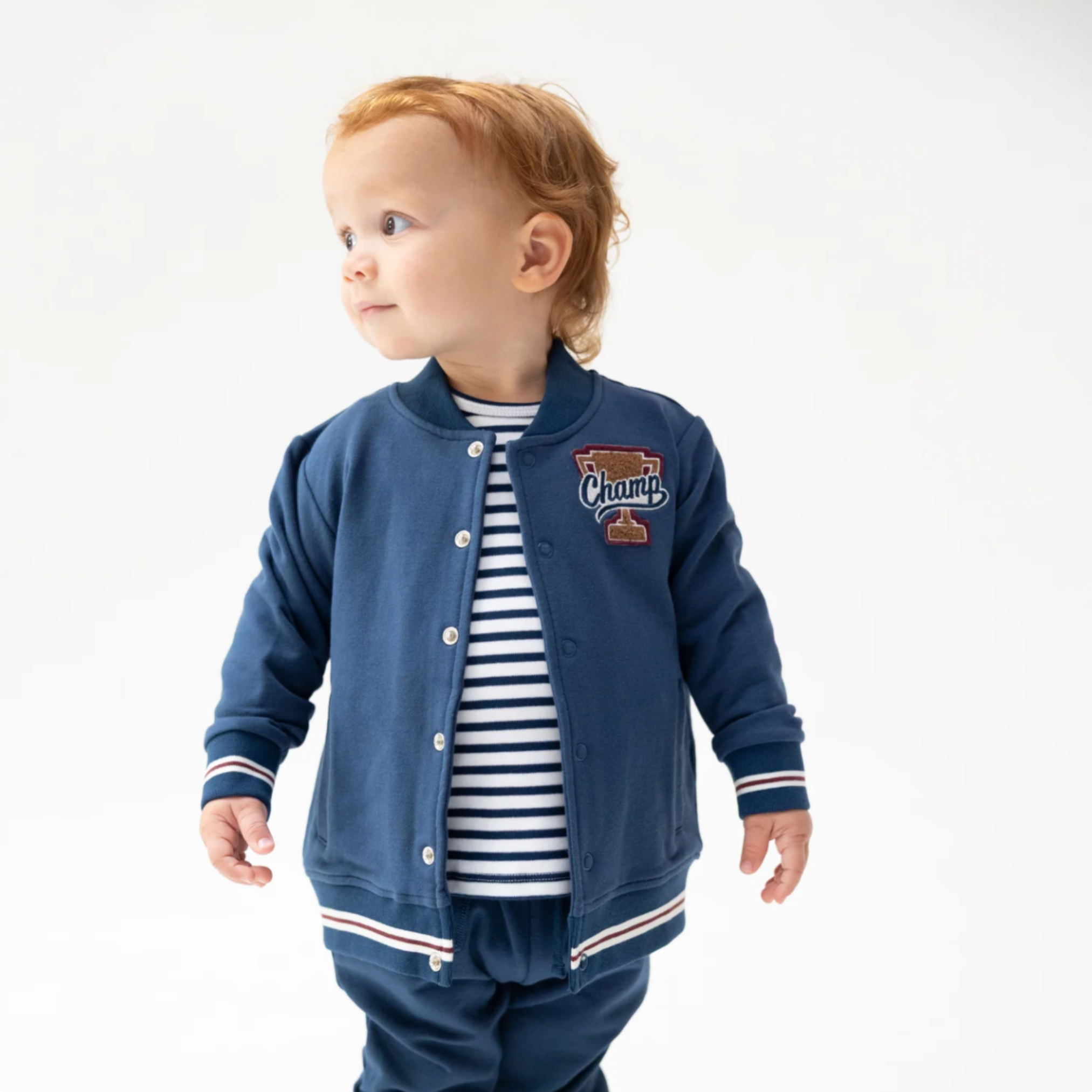 A toddler with red hair wearing a navy blue French Terry varsity jacket over a navy and white striped shirt, paired with matching navy French Terry pants, photographed against a clean white background.