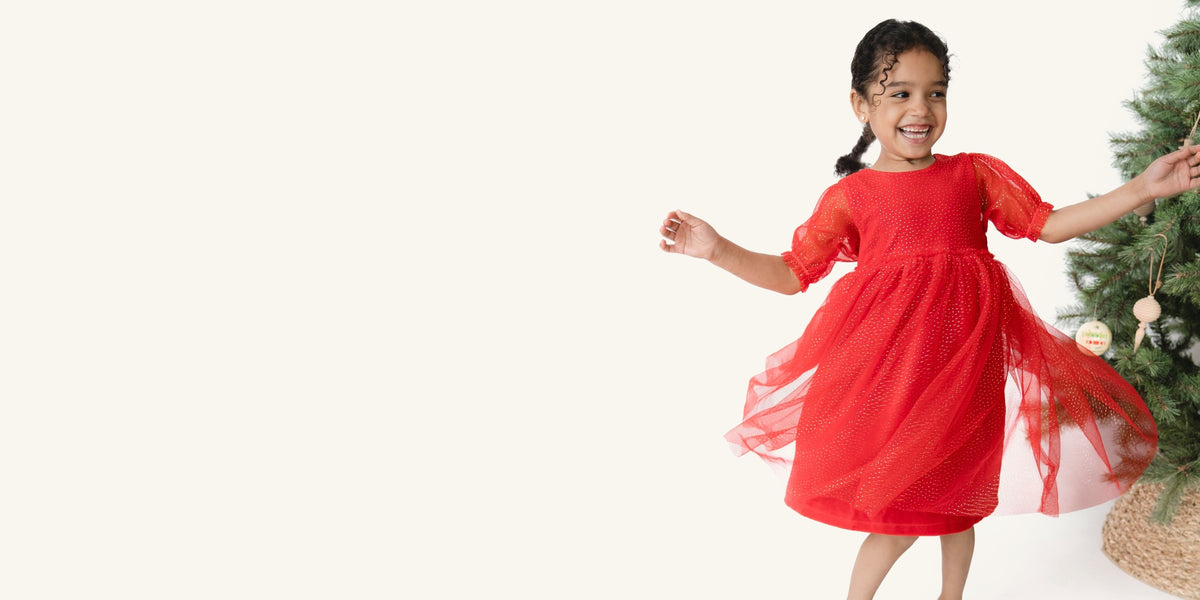 Girl twirls around in a red velour and tulle dress with gold specks. A Christmas tree is to her side.