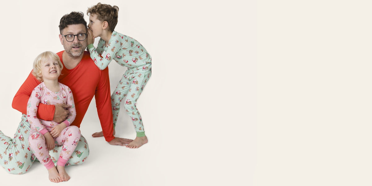 Little boy whispers into dad's ear. Dad has daughter on lap. All wear matching holiday loungewear.