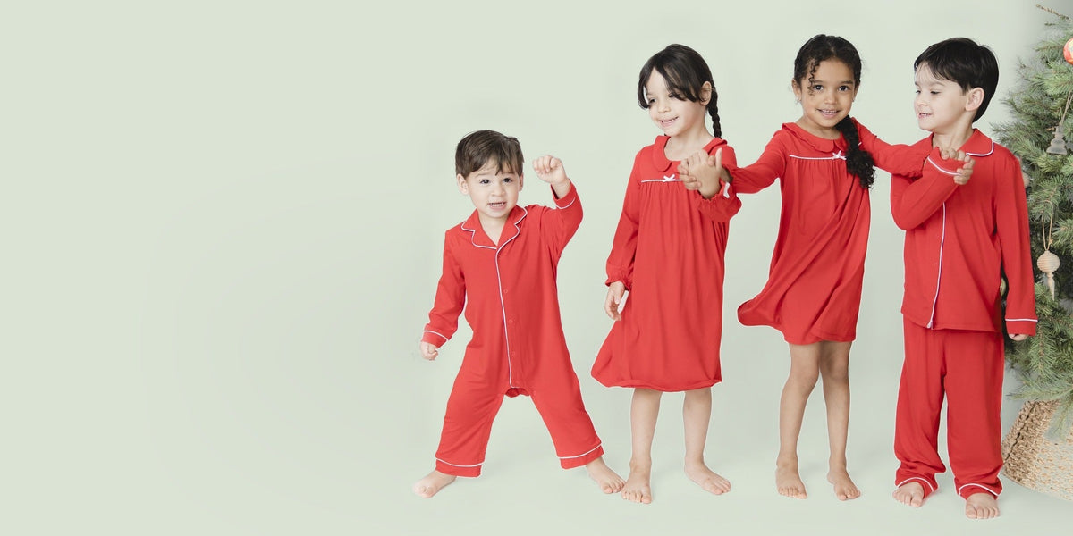 Four young siblings match in the red ribbon holiday loungewear collection. The two girls wear night gowns. The younger boy wear a one piece and the older boy wears a two piece.