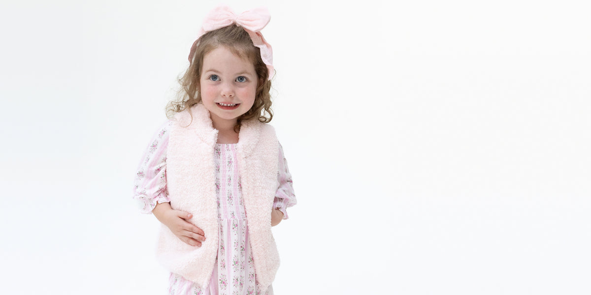 Toddler girl wears a pink striped floral printed bamboo longsleeve dress under a cozy light pink chenille vest and large pink bow on her head.