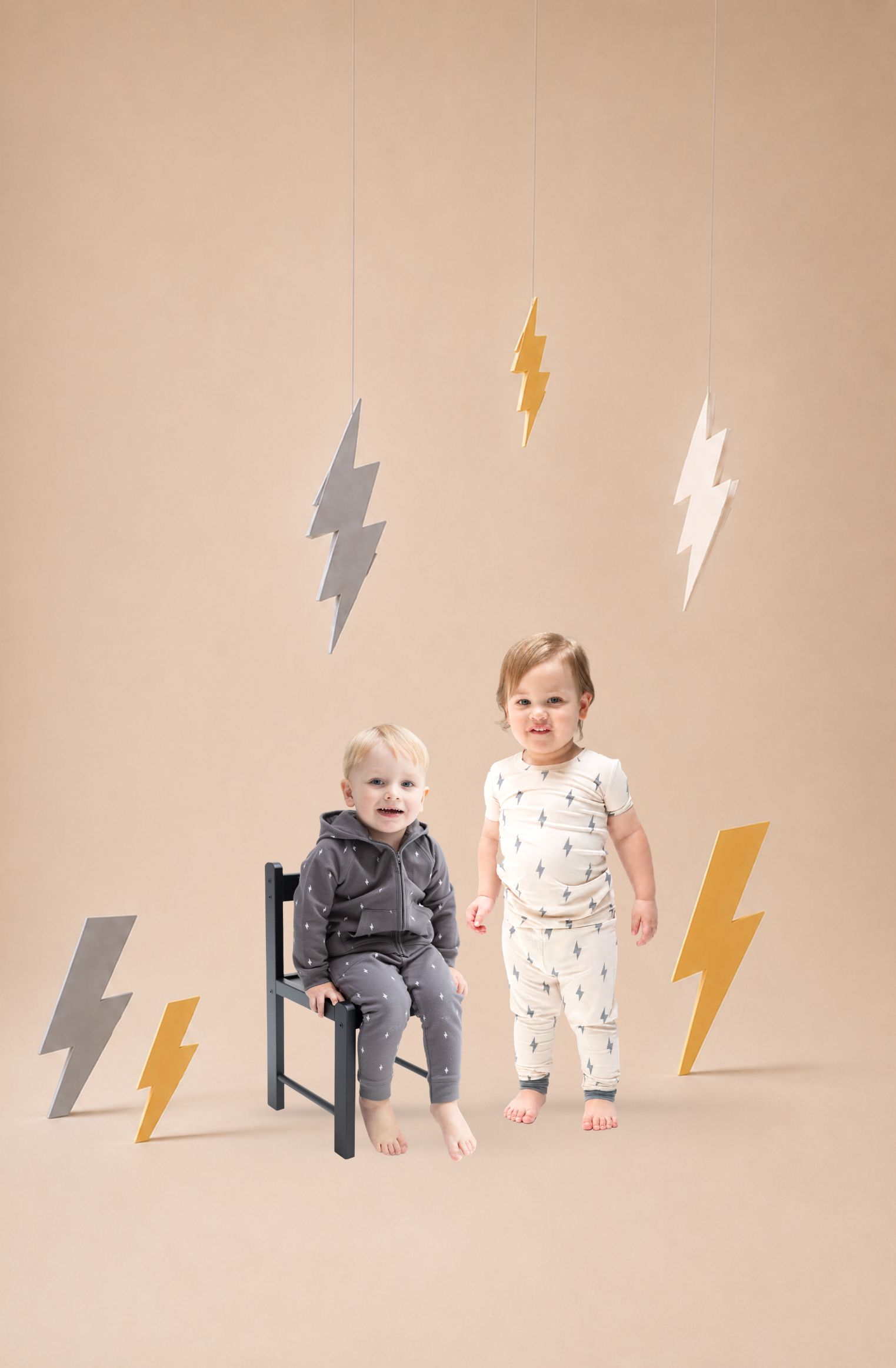 Two toddler boys wear lightening bolt printed outfits on a warm white background with large lightening bolt props around the set.