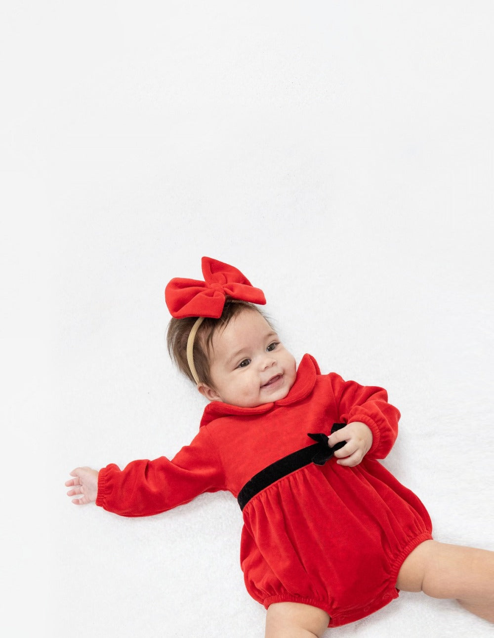 Baby wearing a red velour bubble with a black velvet ribbon at the waist. she also wears a big red velour bow on stretchy headband.
