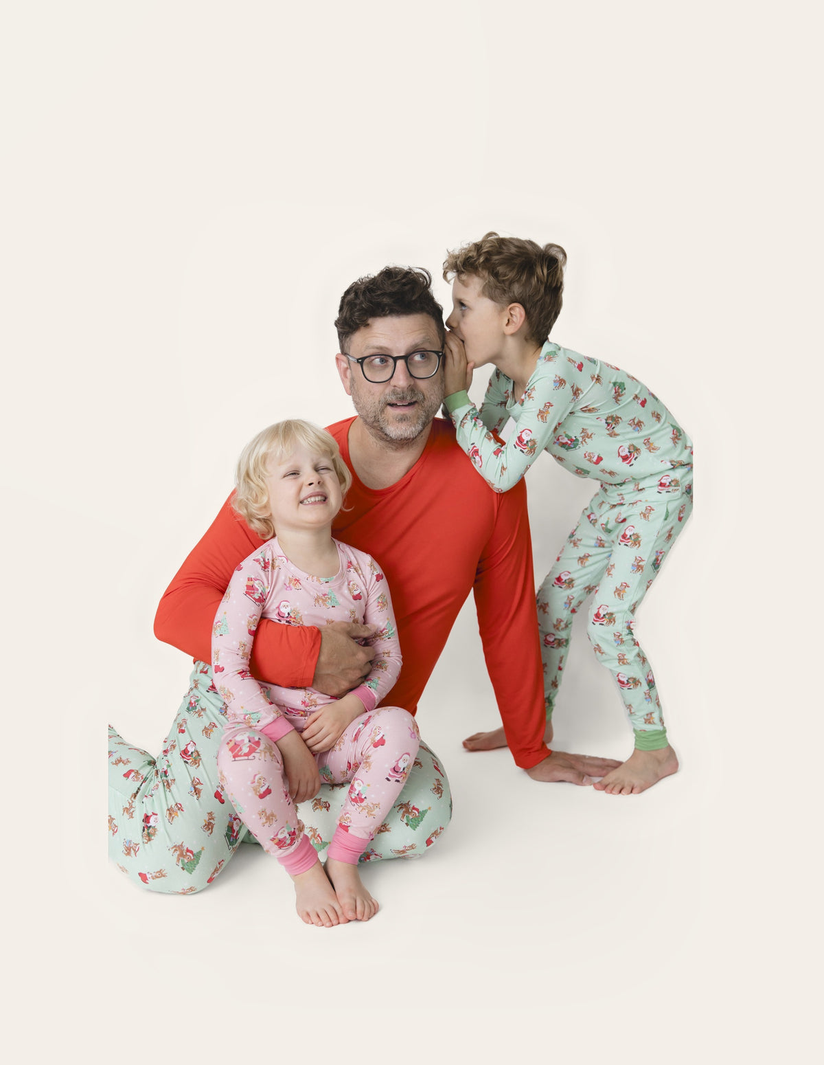 Little boy whispers into dad's ear. Dad has daughter on lap. All wear matching holiday loungewear.