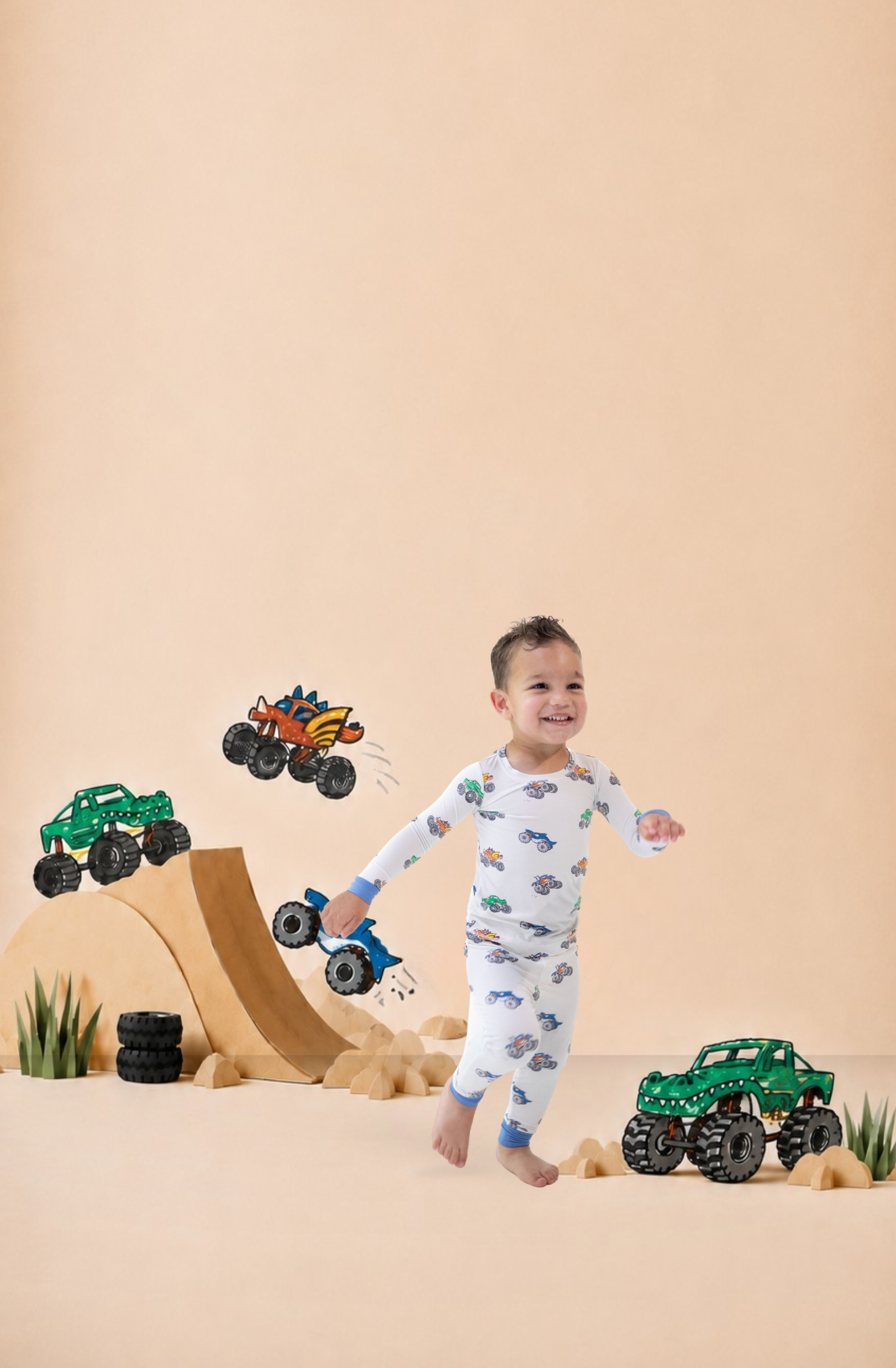 Boy wears pajama set with monster truck print as he runs through a photography set with wooden ramps and overlayed monster truck illustrations taken from the print he is wearing.