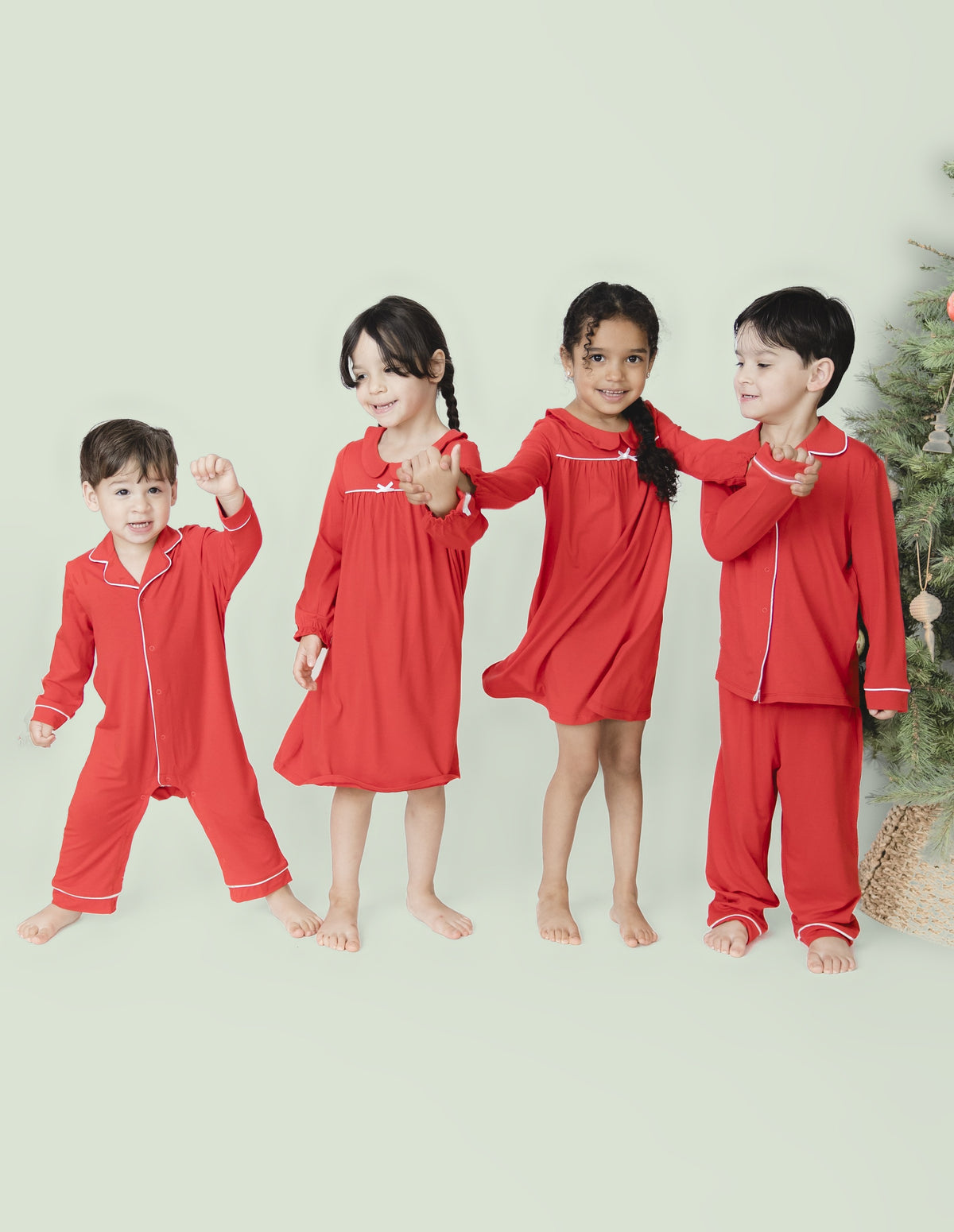 Four young siblings match in the red ribbon holiday loungewear collection. The two girls wear night gowns. The younger boy wear a one piece and the older boy wears a two piece.