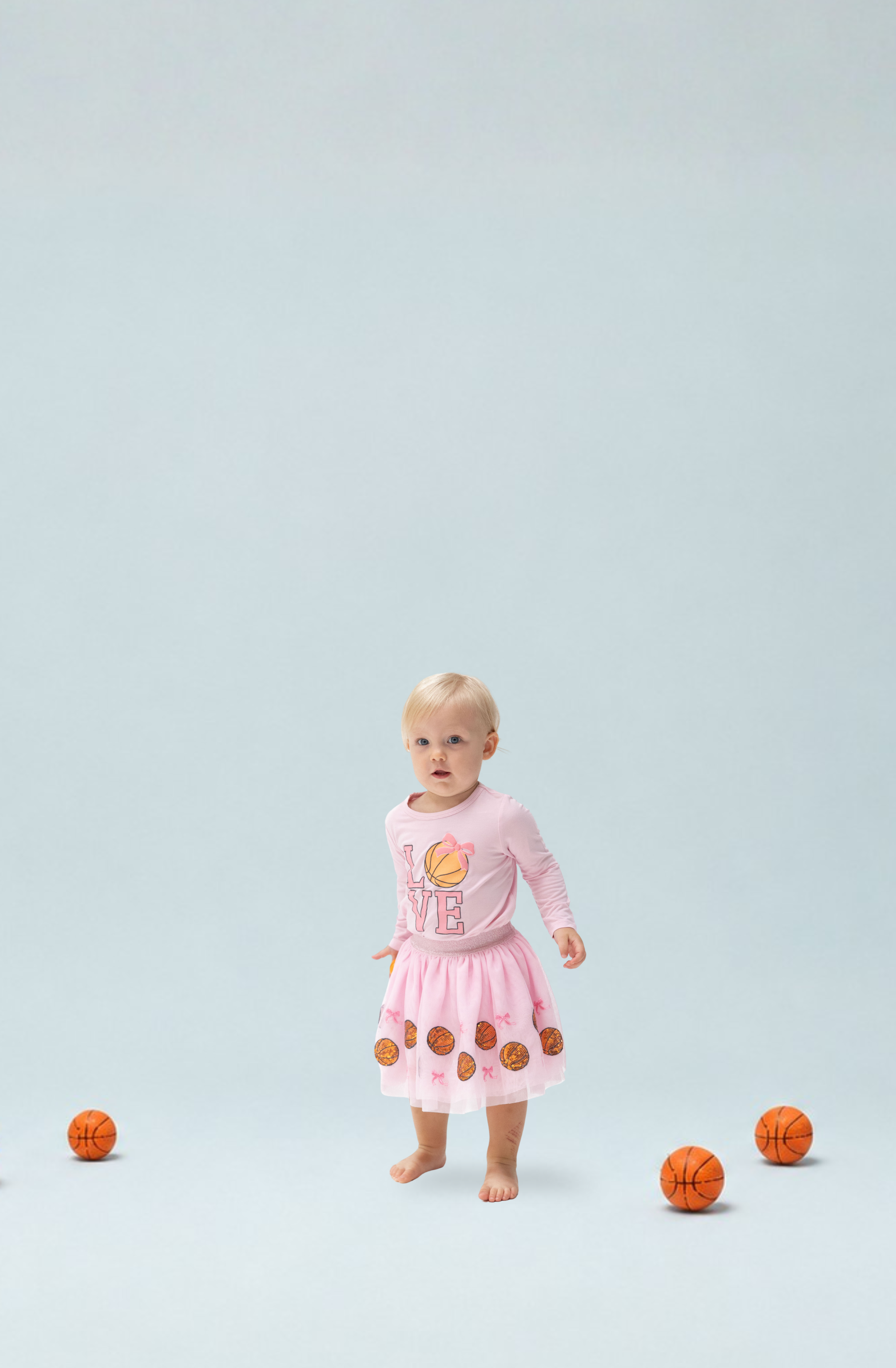 A toddler girls stands barefoot in a light blue studio setting wearing a pink long-sleeve shirt with basketball graphics, and a basketball tutu skirt surrounded by several small orange basketballs.