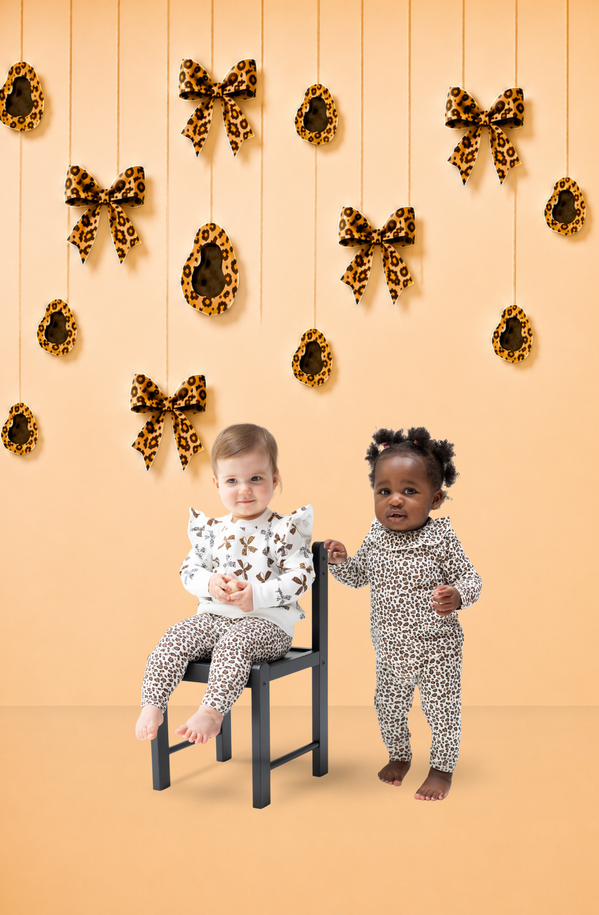 Two toddlers modeling leopard-print outfits. One wears an oversized sweat shirt and leggings and the other wears a one piece with stylized collar. The studio setting is a beige backdrop, hanging leopard bows and spots, and a small black chair.