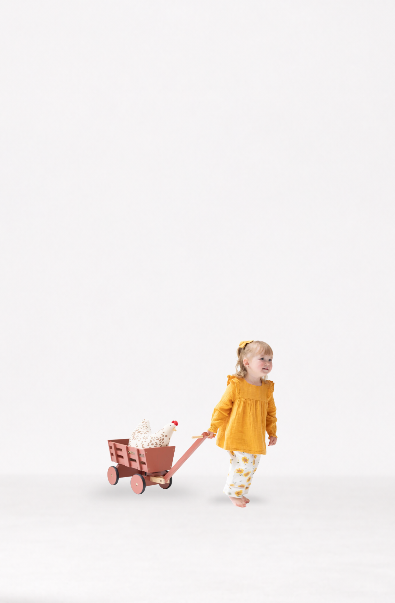 A toddler girl in a muslin bright yellow long-sleeve top and muslin yellow-and-white patterned pants pulls a small wooden wagon with a white stuffed animal chicken, against a seamless white studio background.