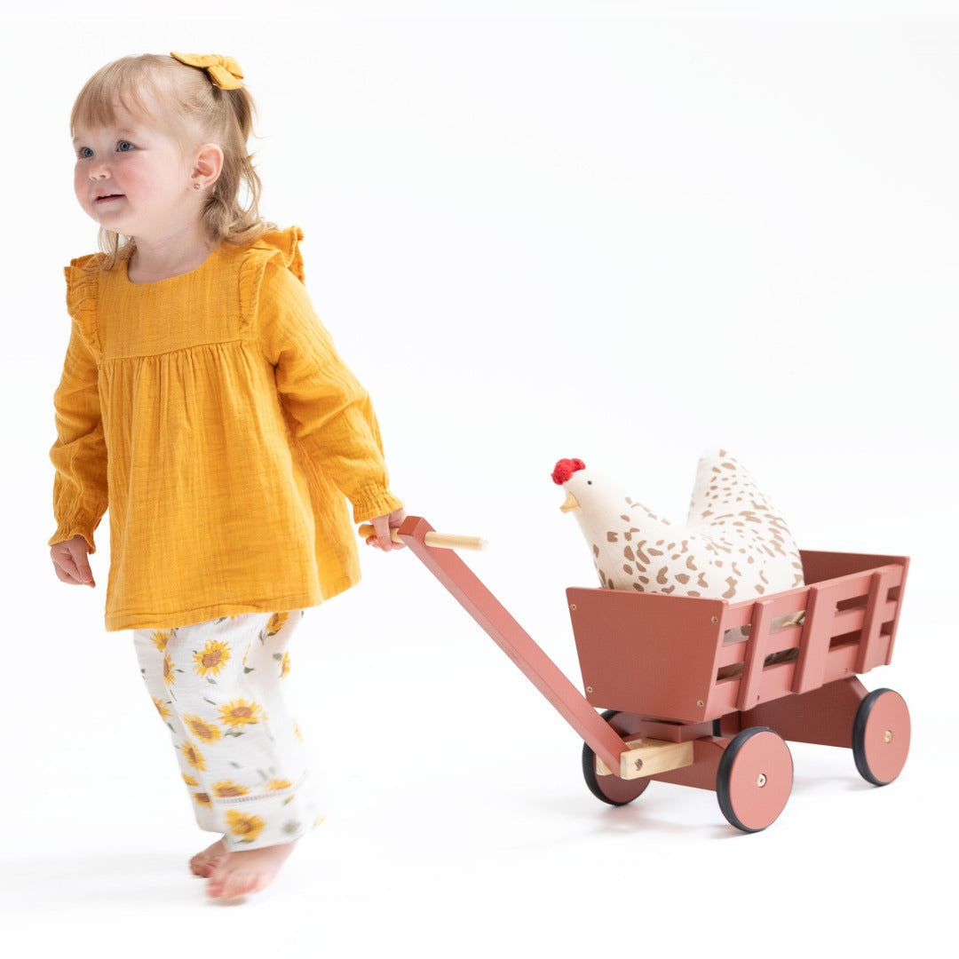 Toddler girl wears a bright sunflower colored tunic top with shoulder ruffles paired with wide legged pants with a sunflower print. Both in muslin. She pulls a red wagon with a toy chicken int he back.