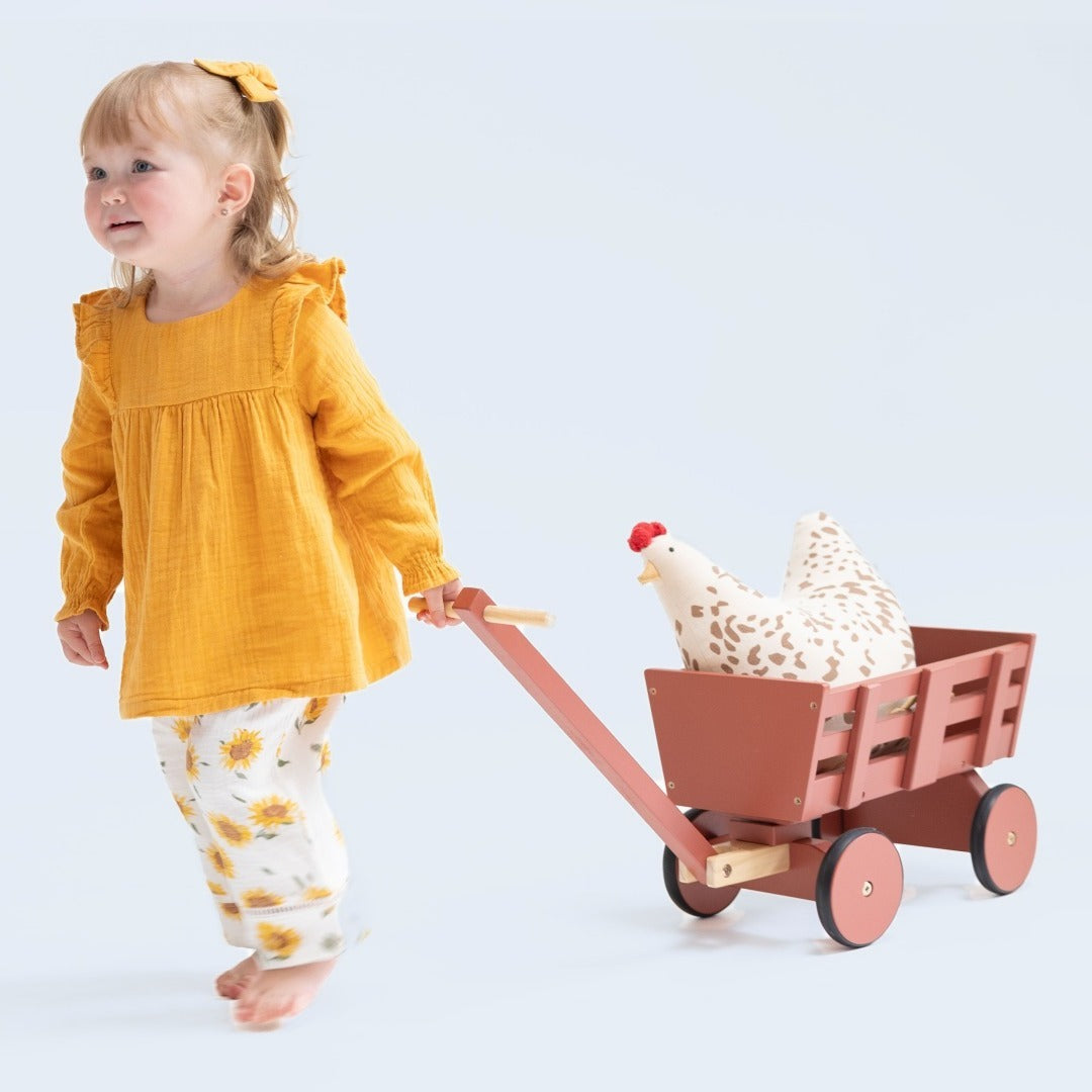 Toddler girl wears a bright sunflower colored tunic top with shoulder ruffles paired with wide legged pants with a sunflower print. Both in muslin. She pulls a red wagon with a toy chicken int he back.