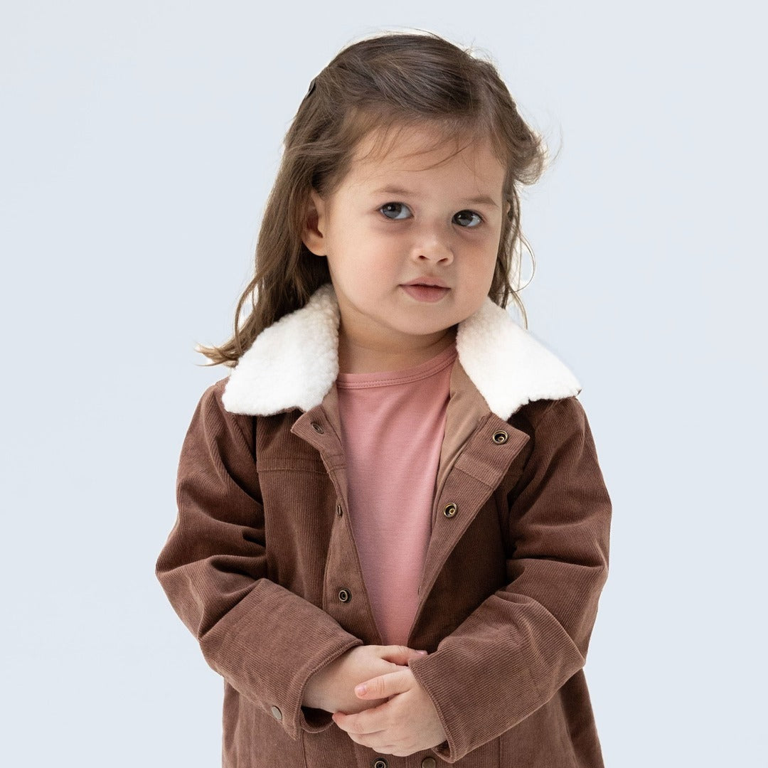 A toddler girl wears the corduroy jacket with white sherpa collar and sweetly looks at the camera.