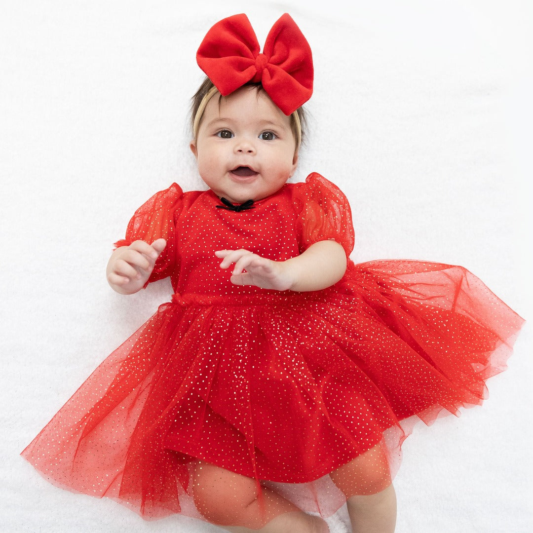 Baby in red velour and tulle dress with gold spec details and small black bow. She also wears a big red velour bow.