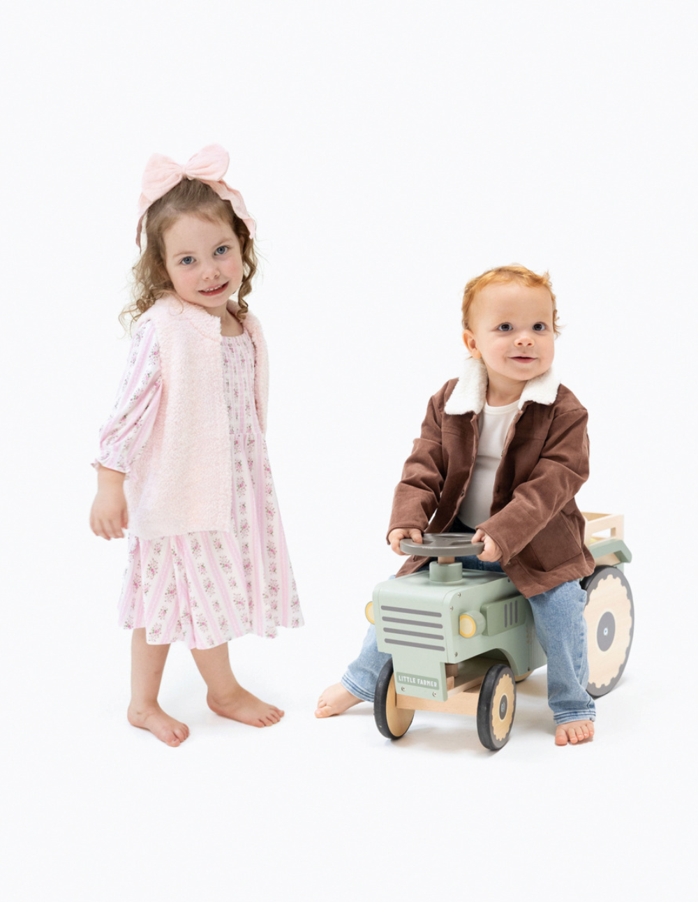 Two young children on a seamless white studio background. On the left, a toddler girl with a pink bow headband wears a light pink long-sleeve dress with a subtle floral pattern and stands barefoot.