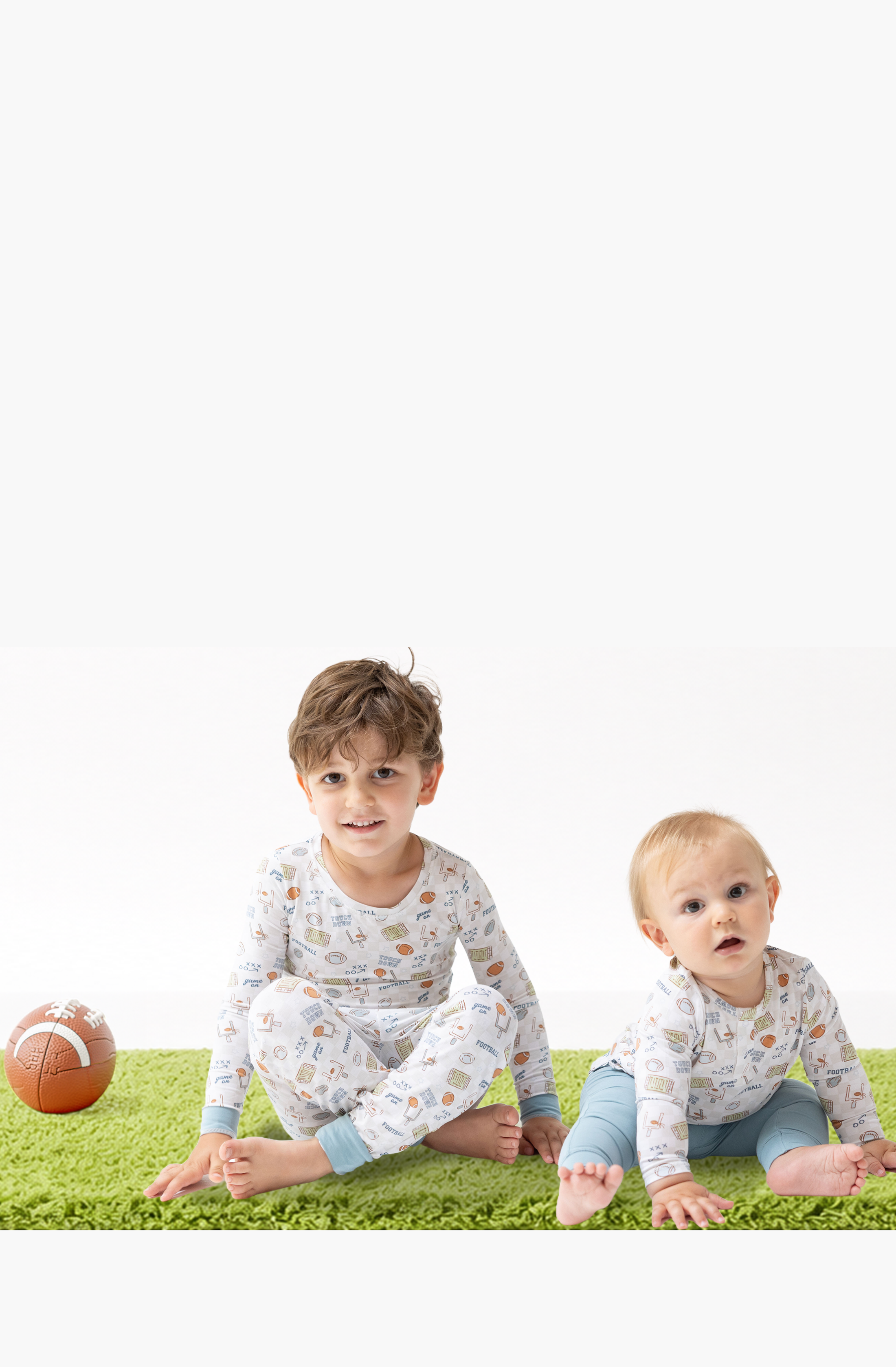 A baby boy and older older brother wearing football printed outfits in soft bamboo fabric, sitting on a bright green rug with a football, against a white studio background.