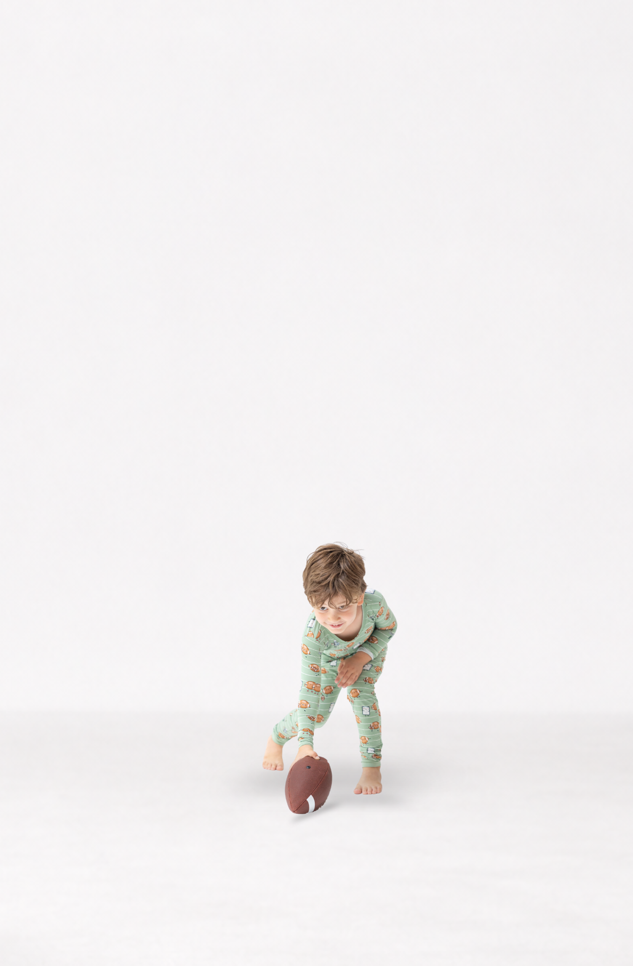 A young child wearing a football patterned pajama set is barefoot, reaching for a brown football on a seamless white studio background.