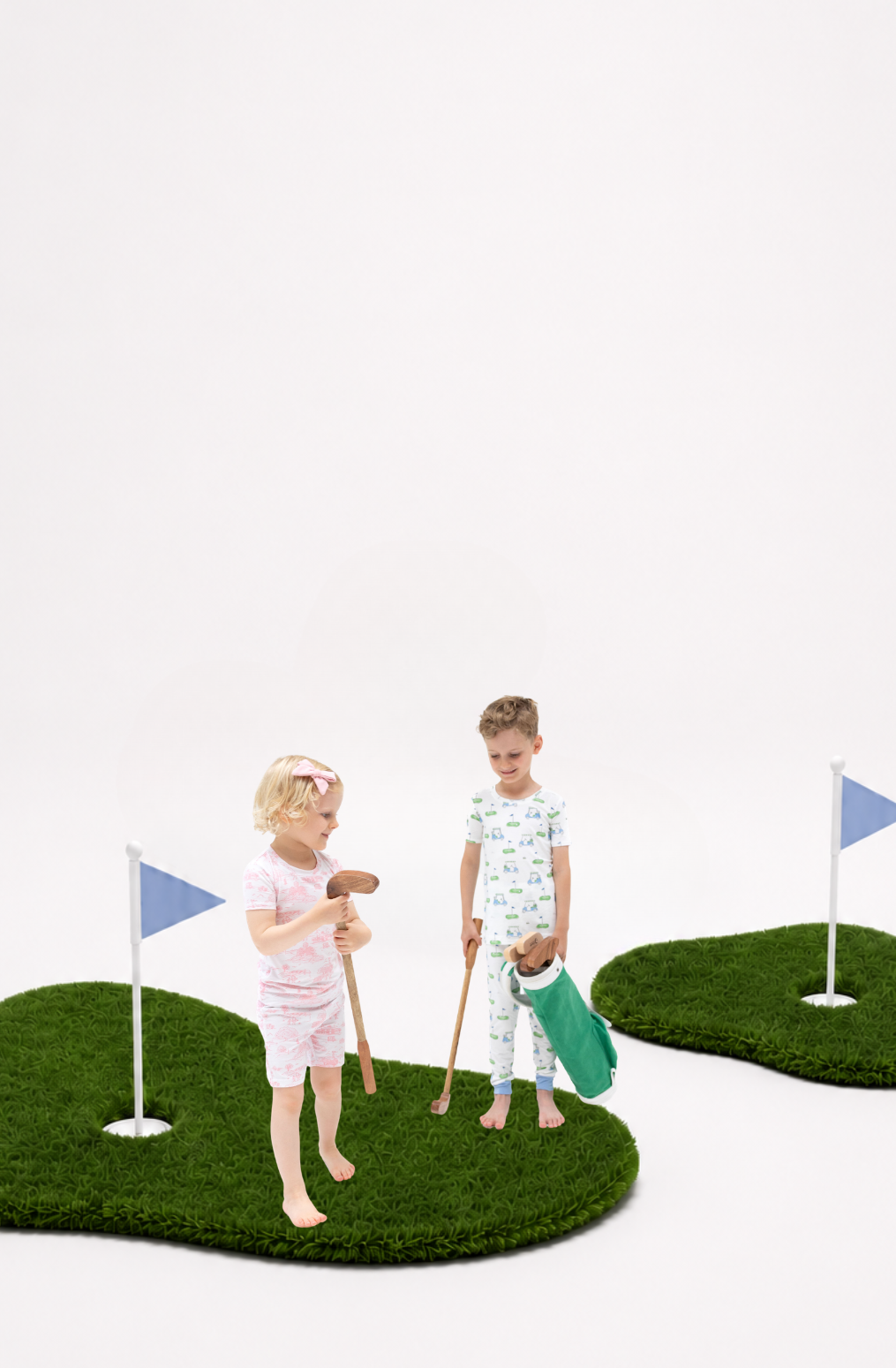 Two children wearing golf printed pajamas playing on small artificial grass golf greens with golf clubs and flags against a plain white background.