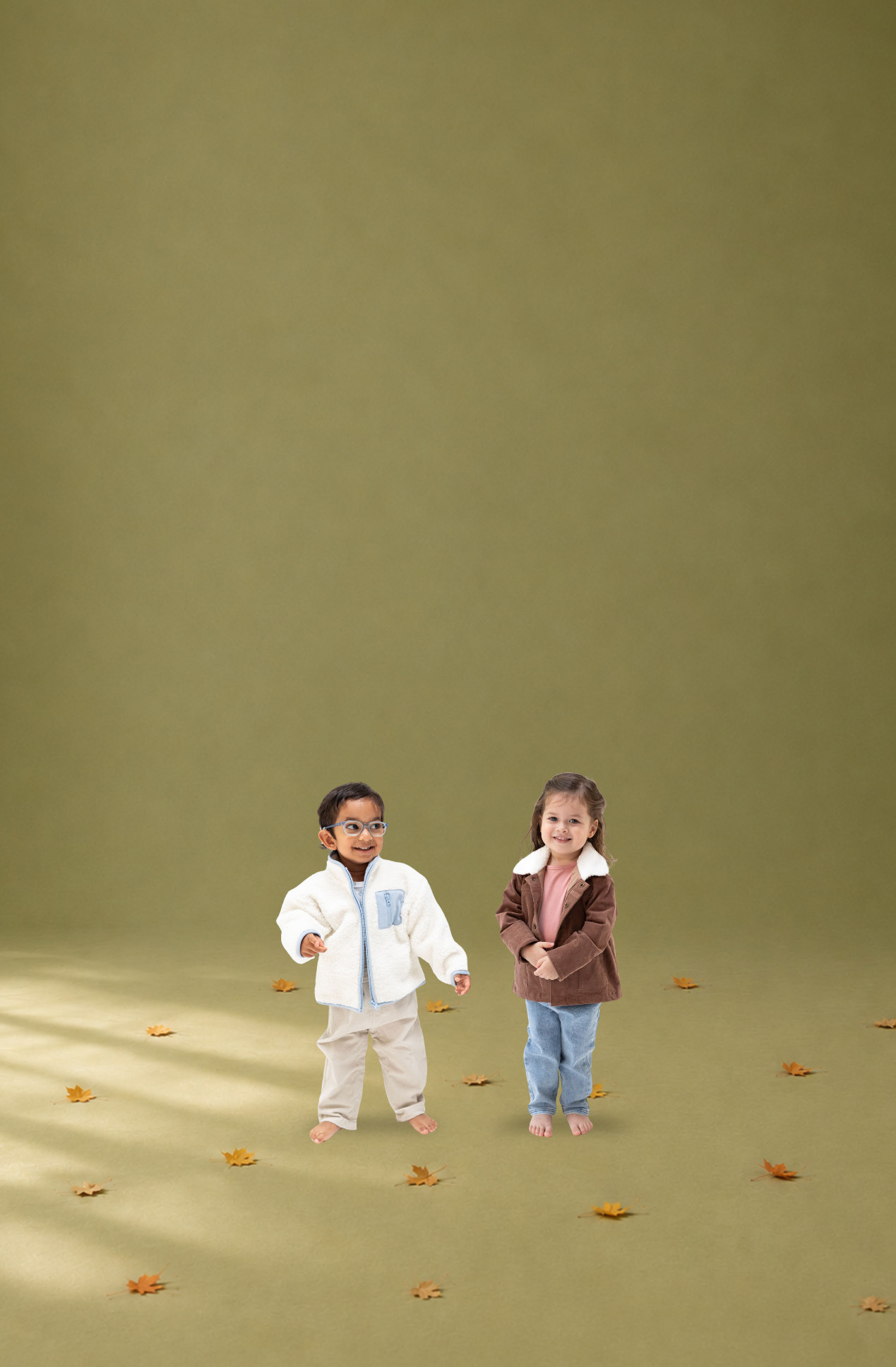 Two young children standing barefoot on a solid olive green studio floor, surrounded by scattered autumn leaves. The child on the left wears a white chenille jacket with a light blue chest panel and beige pants, while the child on the right wears a brown corduroy jacket with a white sherpa collar, a pink top, and blue jeans.