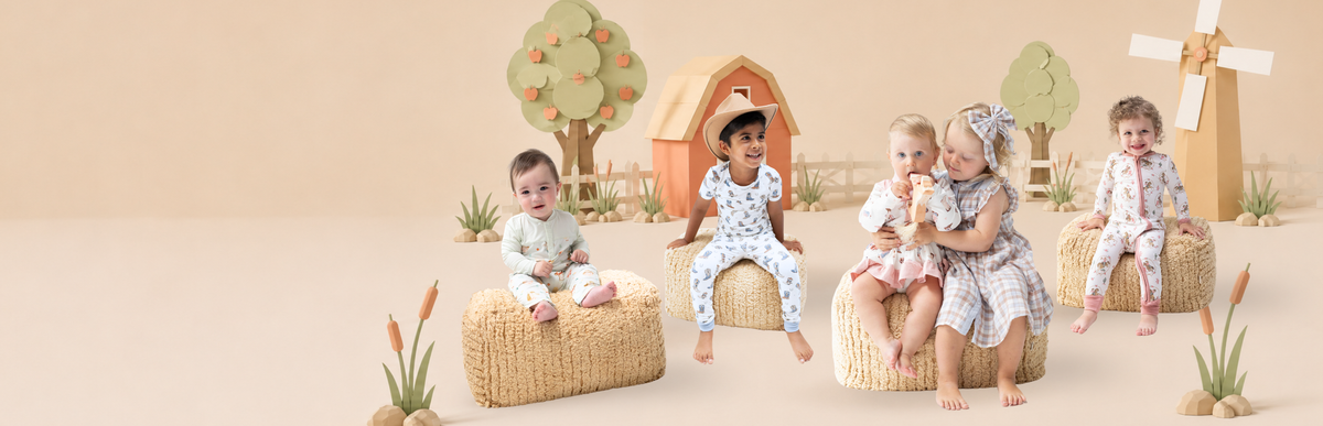 Five toddlers wearing pieces from the Angel Dear Fall Farm collection are posed on woven straw stools in a whimsical, rustic studio set with a small wooden house, trees, and a windmill.