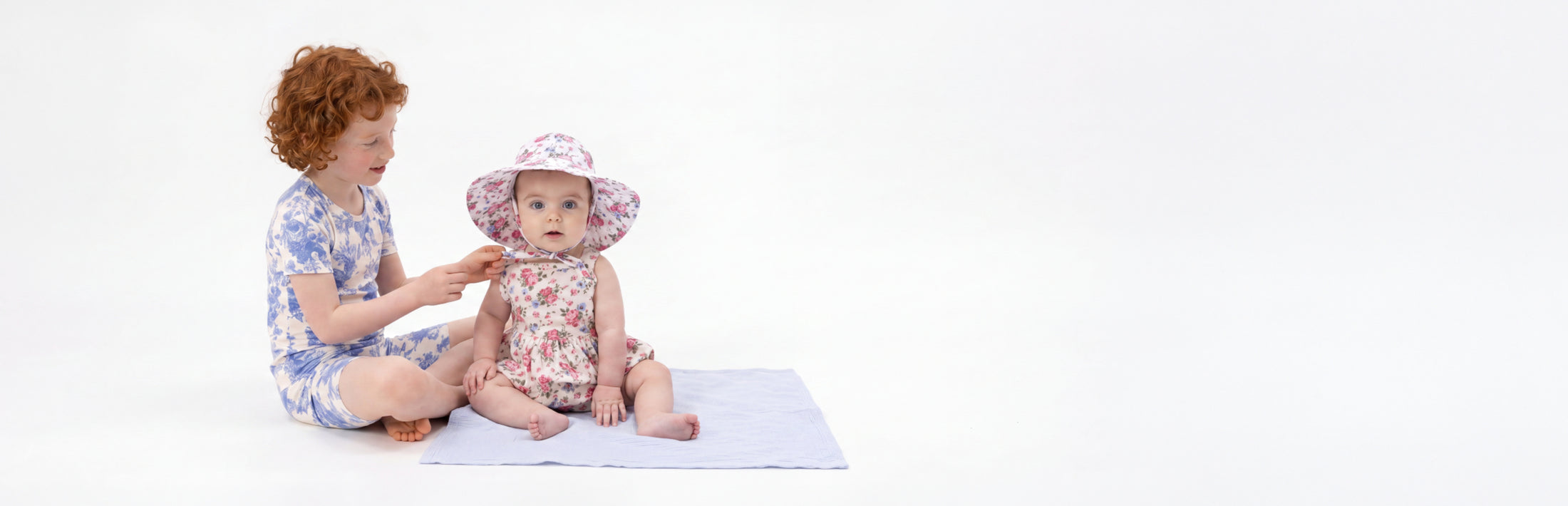 An older sister helps to tie her baby sister's sunhat. Both children wear red, white and blue floral printed outfits.