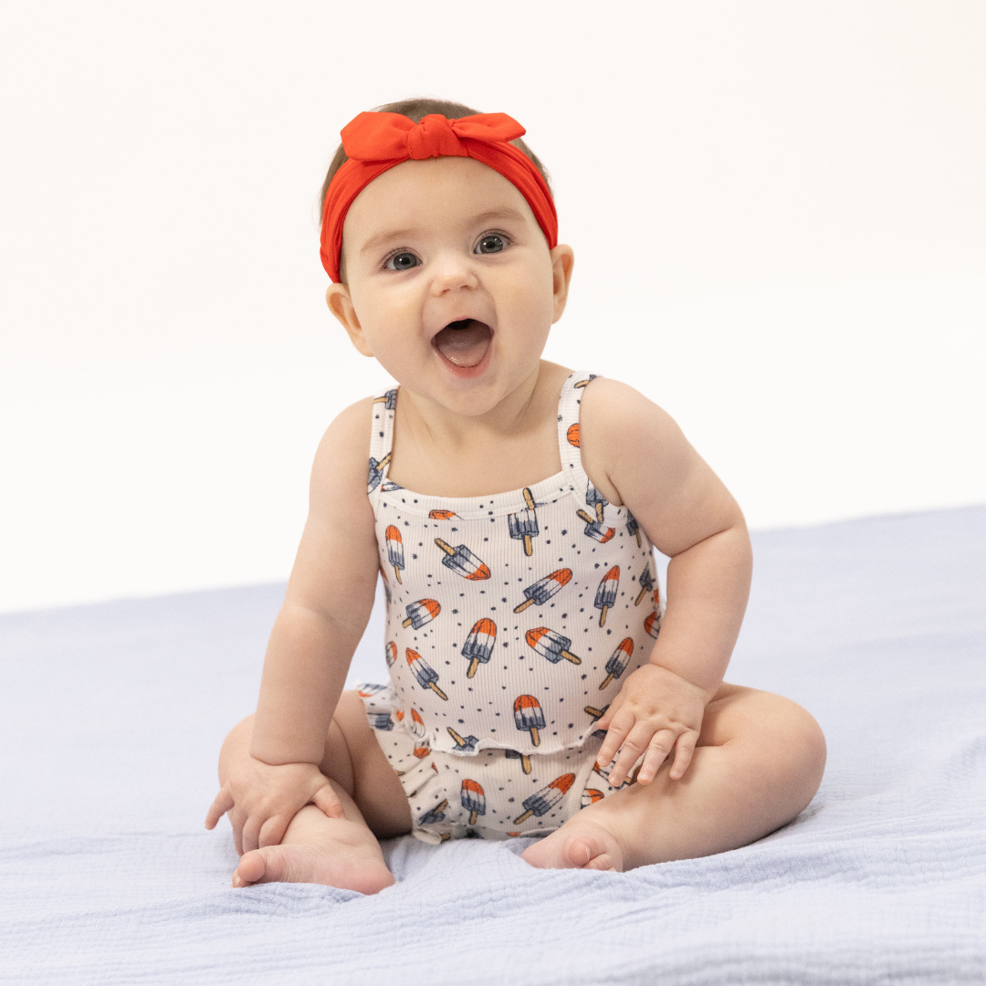 Baby girl wears a Fourth of July themed outfit featuring Astro Pops and a red headband.