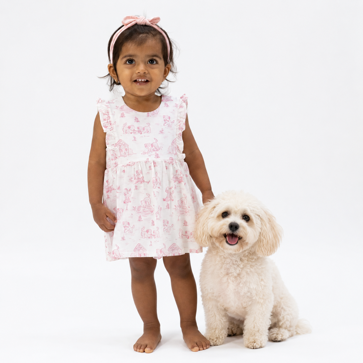 Young girl in a dog printed toile dress standing next to a small white dog on a white background