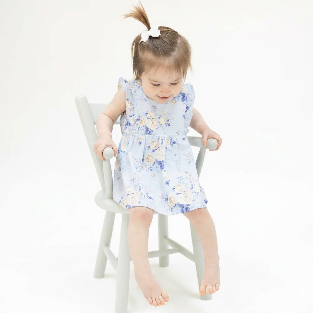 A toddler girl with a high ponytail sits on a light gray wooden chair, wearing a light blue sleeveless dress with a blue and beige floral pattern.